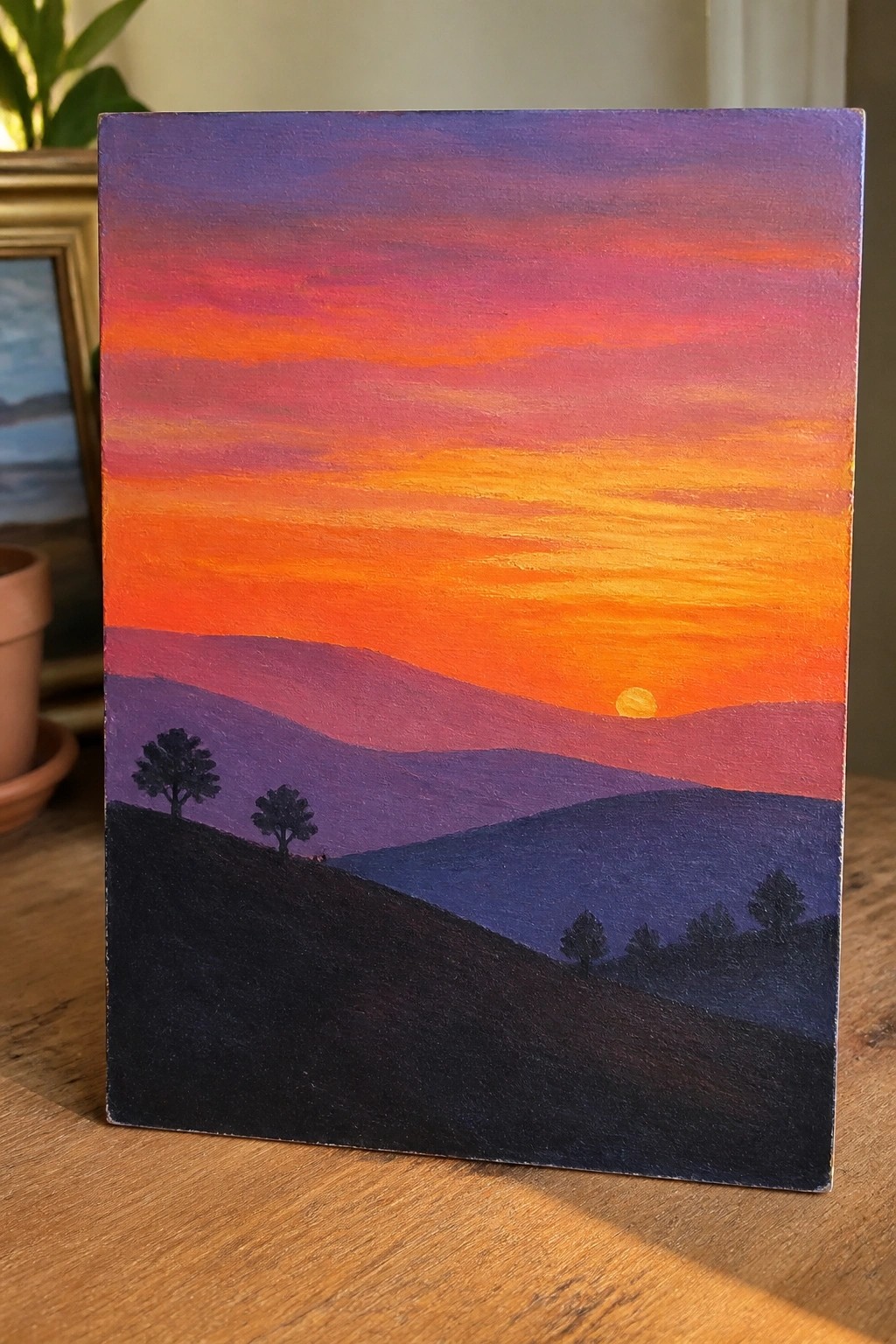 Vibrant sunset painting with layered purple hills, silhouetted trees, and glowing orange sky.