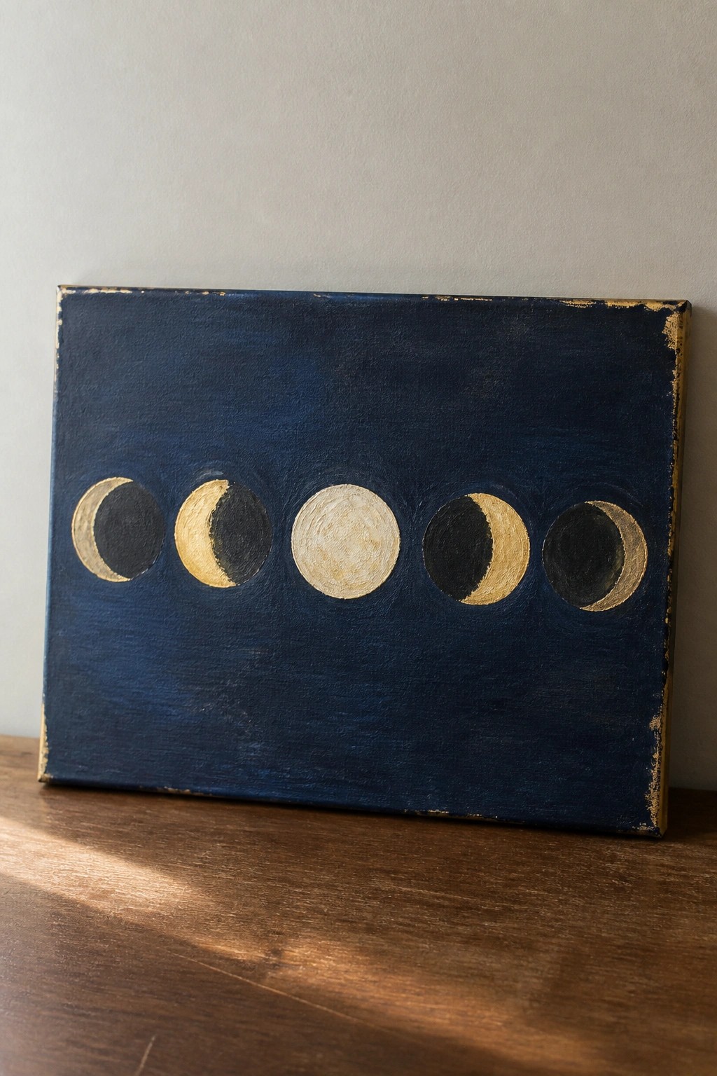 Moon phase painting with gold crescents on navy canvas resting on wooden surface against wall.