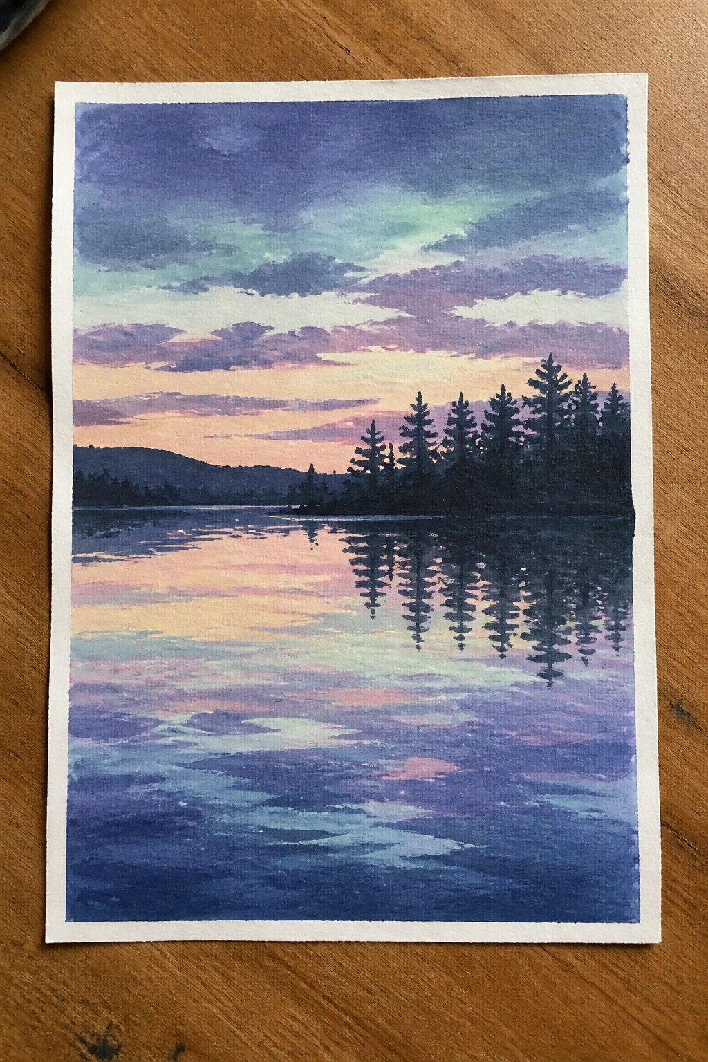 Watercolor painting of a sunset lake with silhouetted pines and vibrant sky reflections.