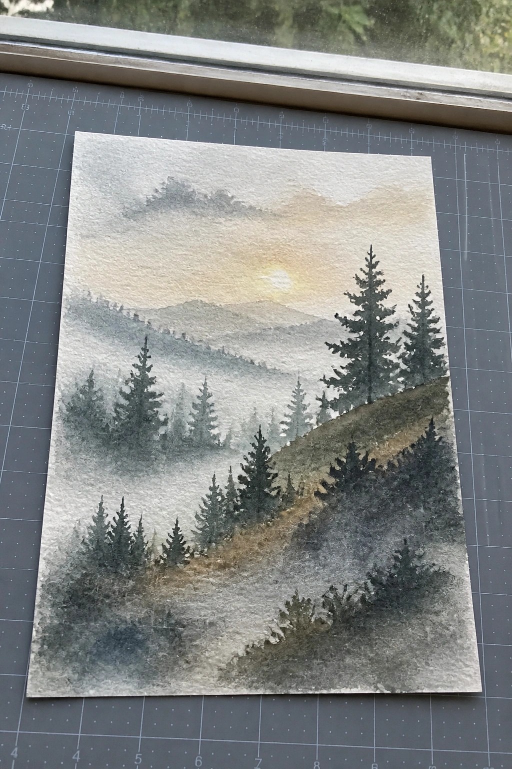Misty watercolor mountains with dark pine trees and hazy golden sunset glow.