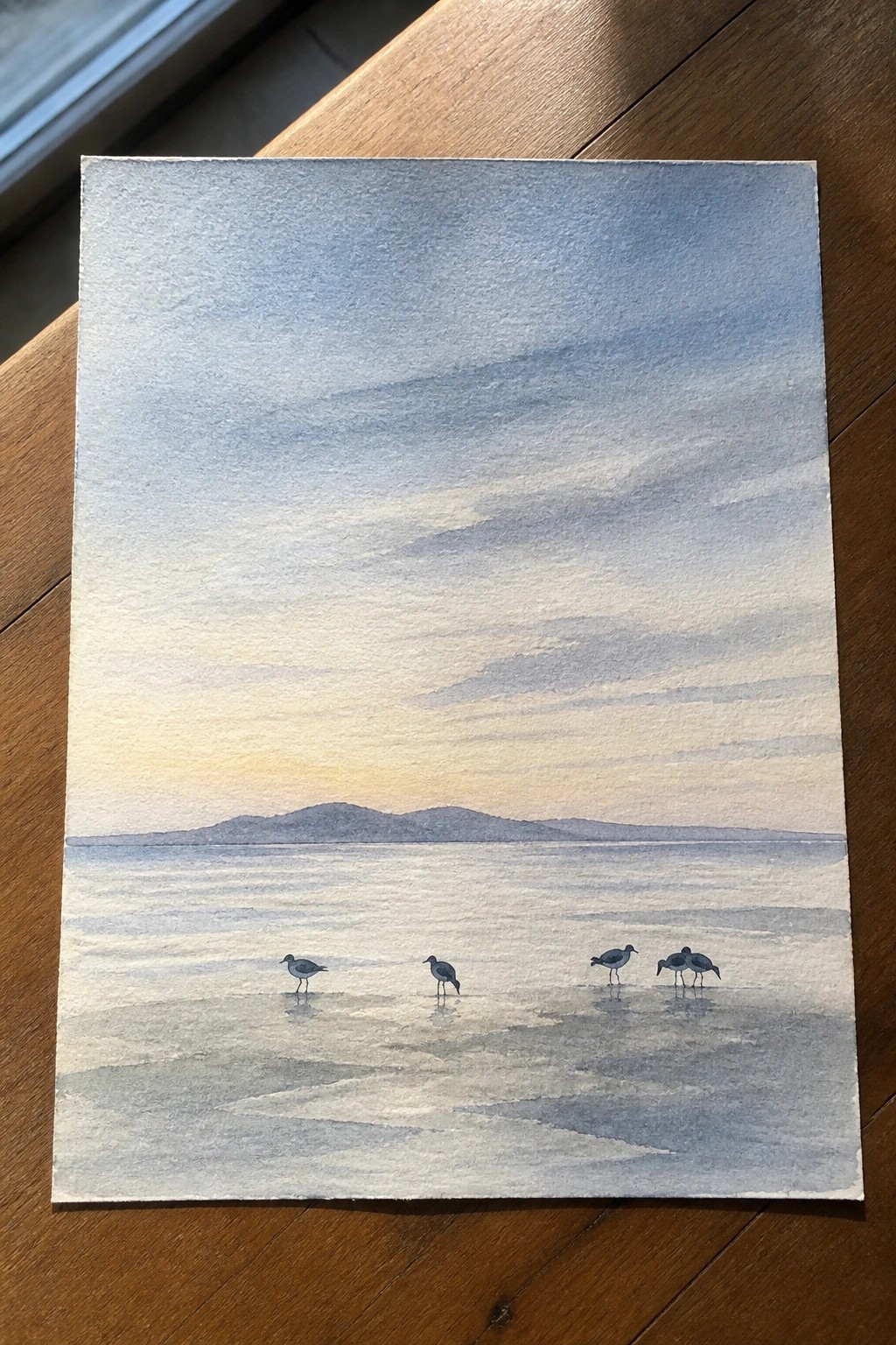 A watercolor painting shows a calm expanse of water with distant hills under a soft gradient sky and four small birds standing on the wet foreground shore.