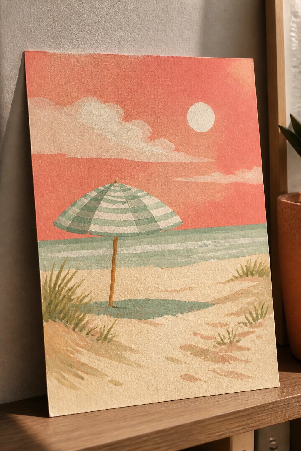 Watercolor painting of a green and white striped beach umbrella on sandy dunes with ocean waves and a red-orange sunset sky featuring a white sun and clouds.