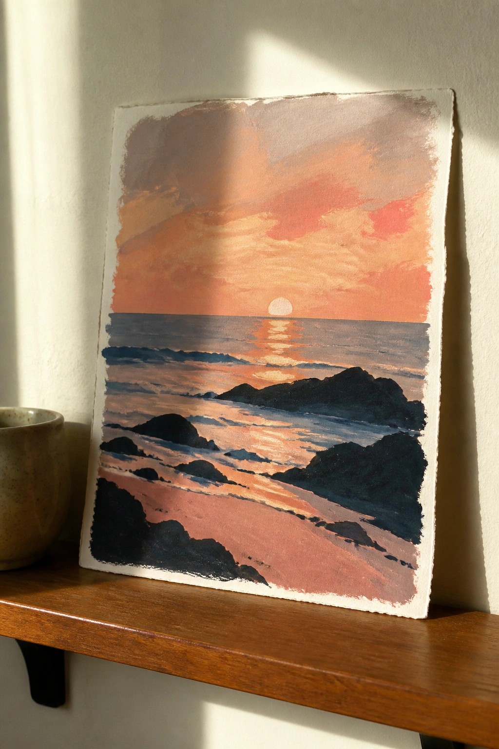 Orange sunset seascape painting on wooden shelf beside speckled ceramic cup