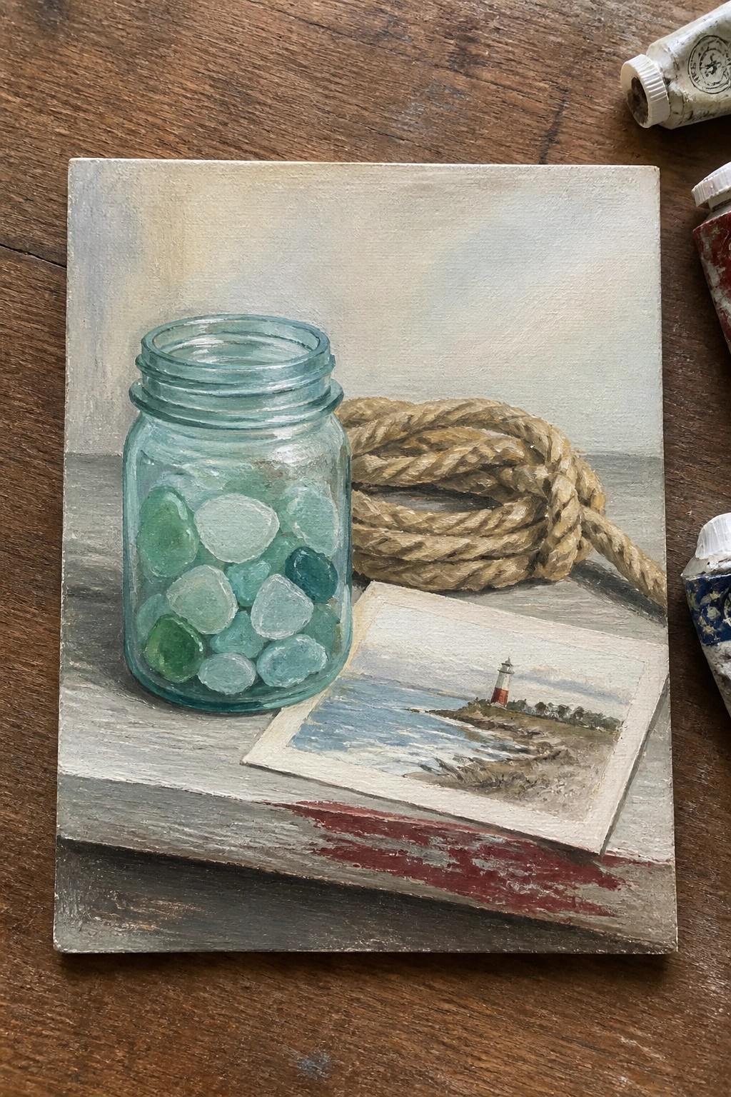 A small oil painting shows a glass jar filled with green and blue sea glass next to a thick rope and a lighthouse postcard on a wooden surface.