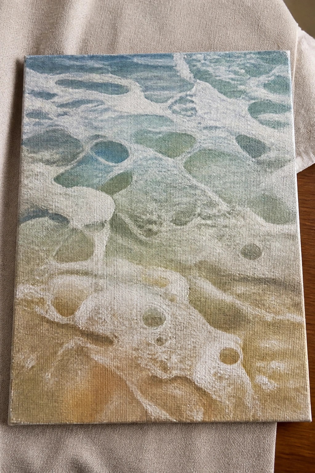 Canvas painting of foamy ocean waves in blues, greens, and white on beige fabric.