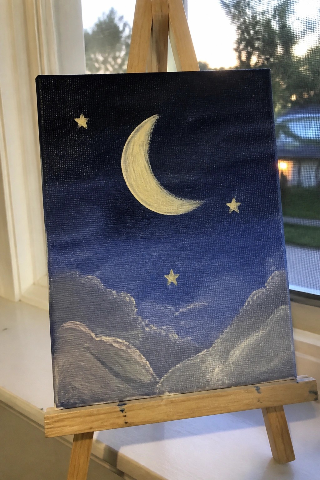 Canvas on wooden easel shows yellow crescent moon, stars, and clouds in blue night sky.