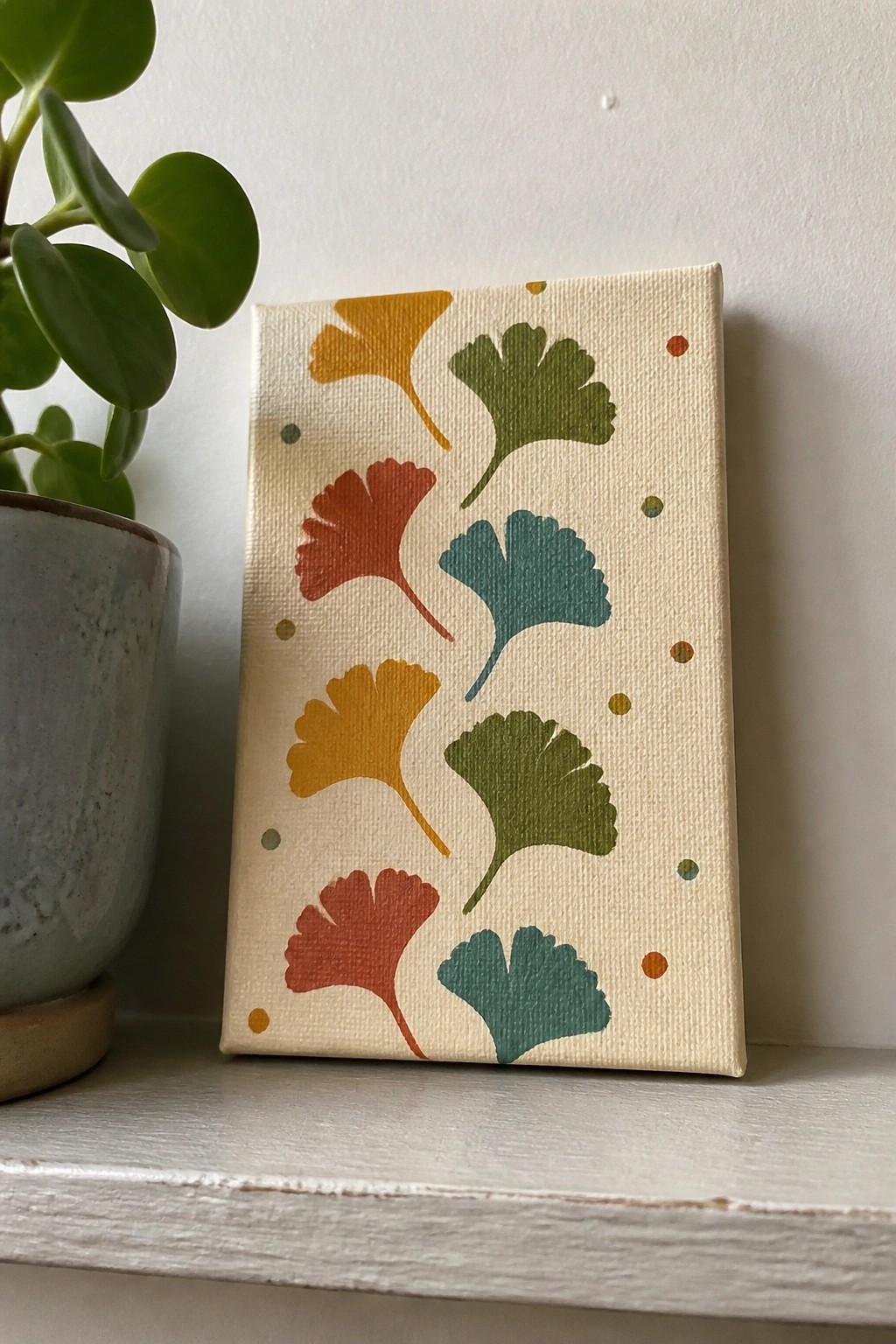 Colorful ginkgo leaf painting on small canvas beside potted plant on white shelf