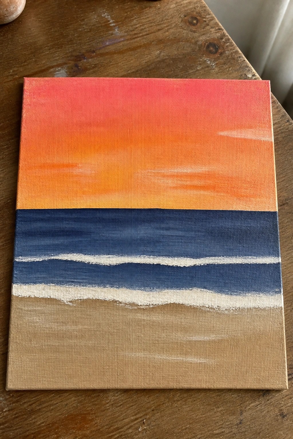 Acrylic painting of red-orange sunset over blue ocean with white waves and brown sand.