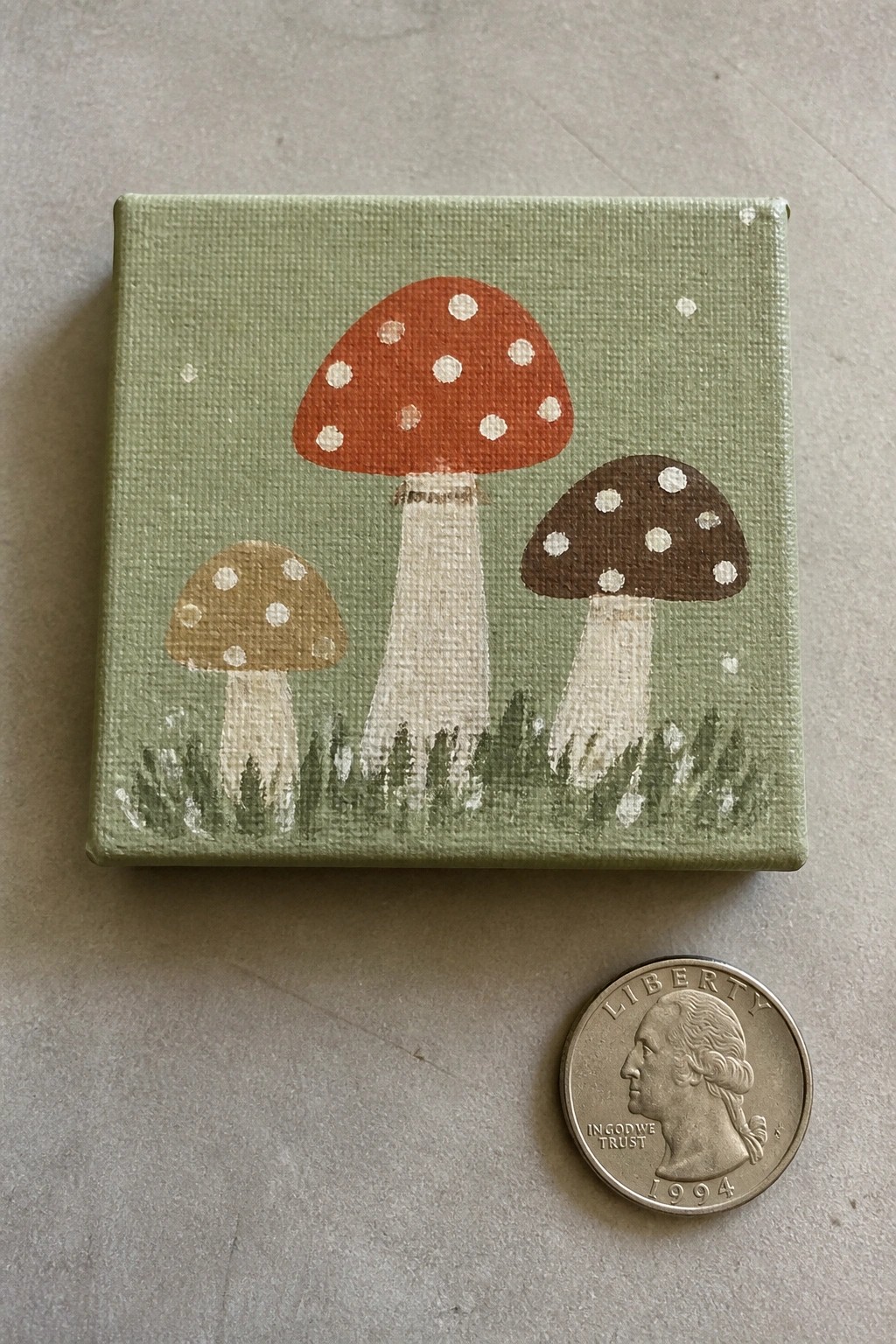 Mini green canvas painting of three spotted mushrooms beside a 1994 quarter.
