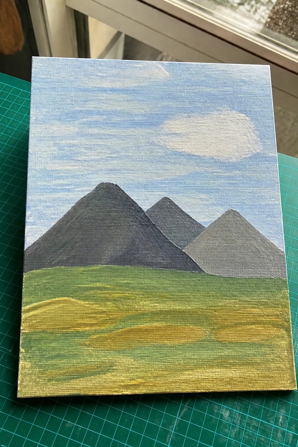 Acrylic painting on canvas of three dark mountains under blue sky with green field
