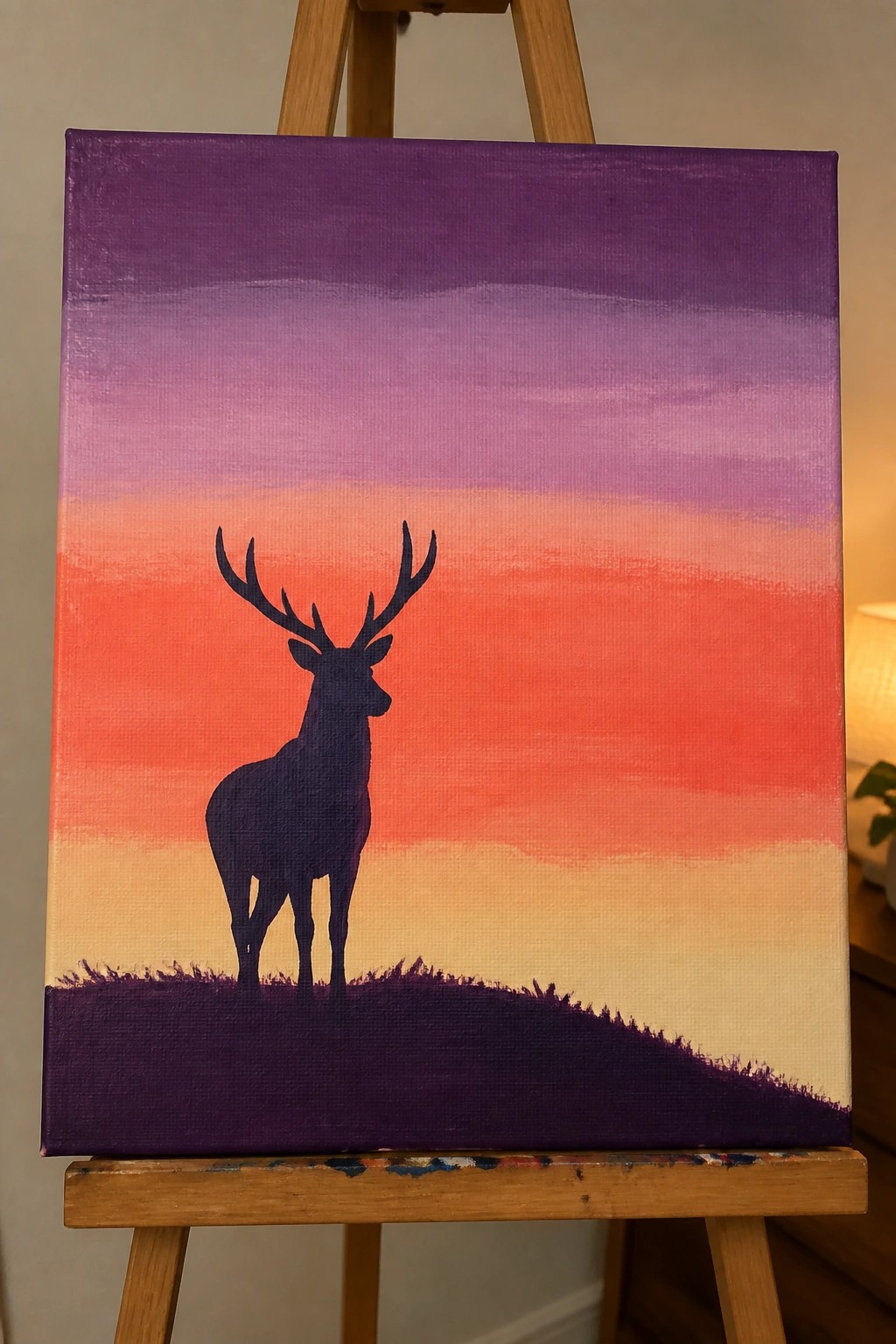 Deer silhouette with antlers on dark hill under purple-orange gradient sky on easel.