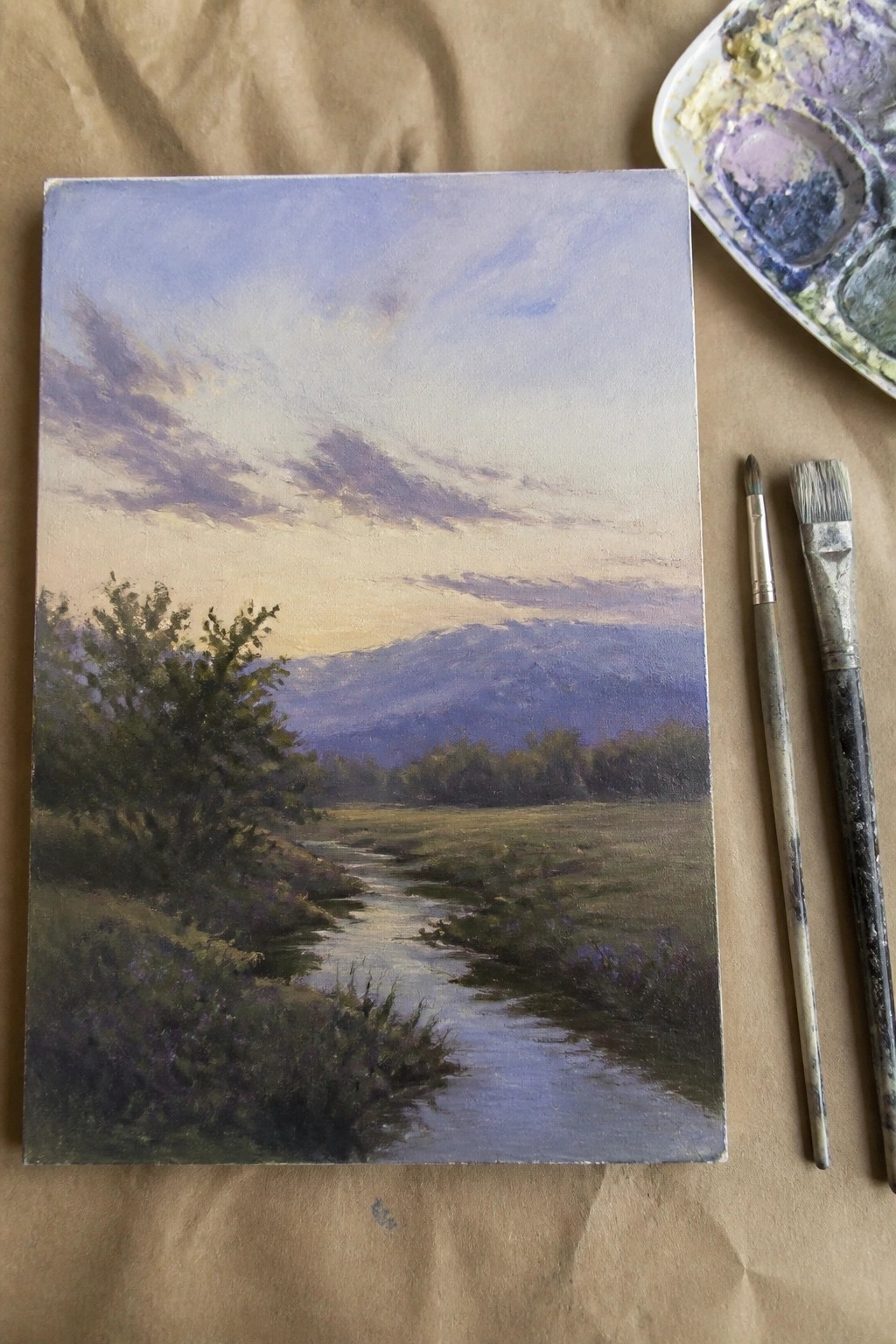 Canvas landscape of winding stream, mountains, trees beside palette and brushes