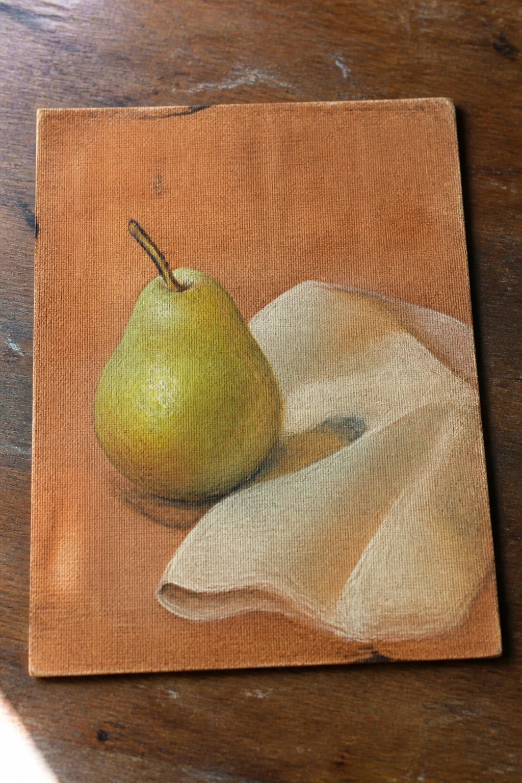 Still-life painting of green pear beside draped white cloth on brown canvas