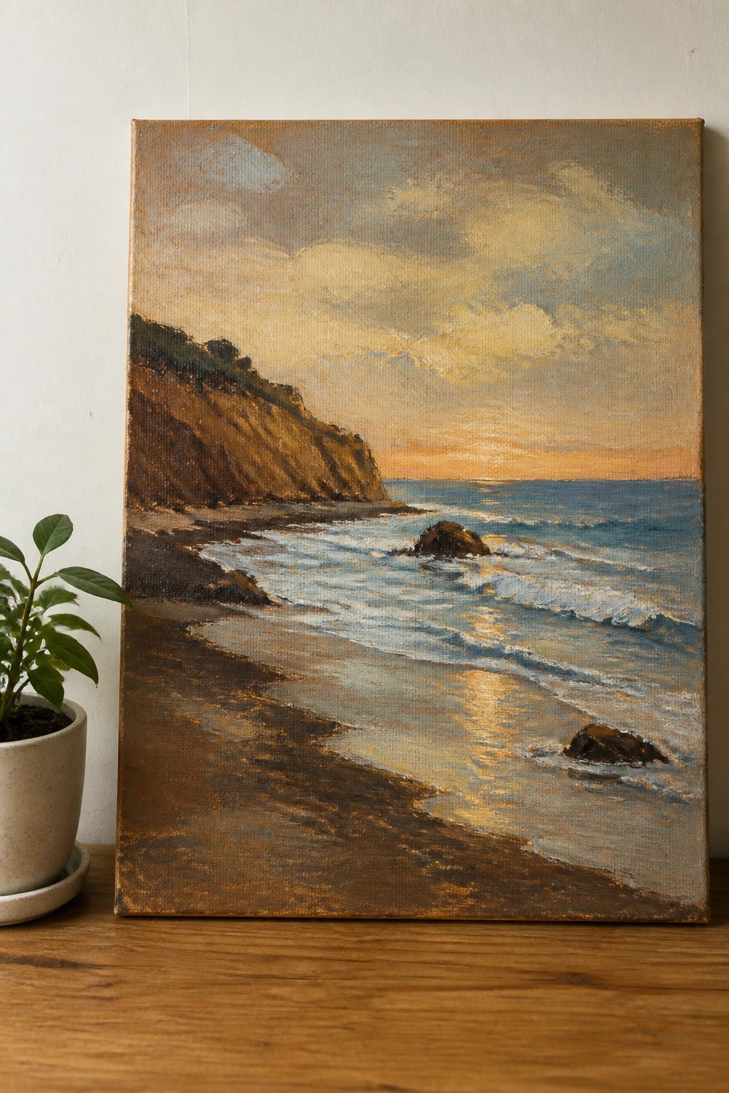 Oil painting of sunset beach with cliffs, waves, and rocks beside potted plant on wood.