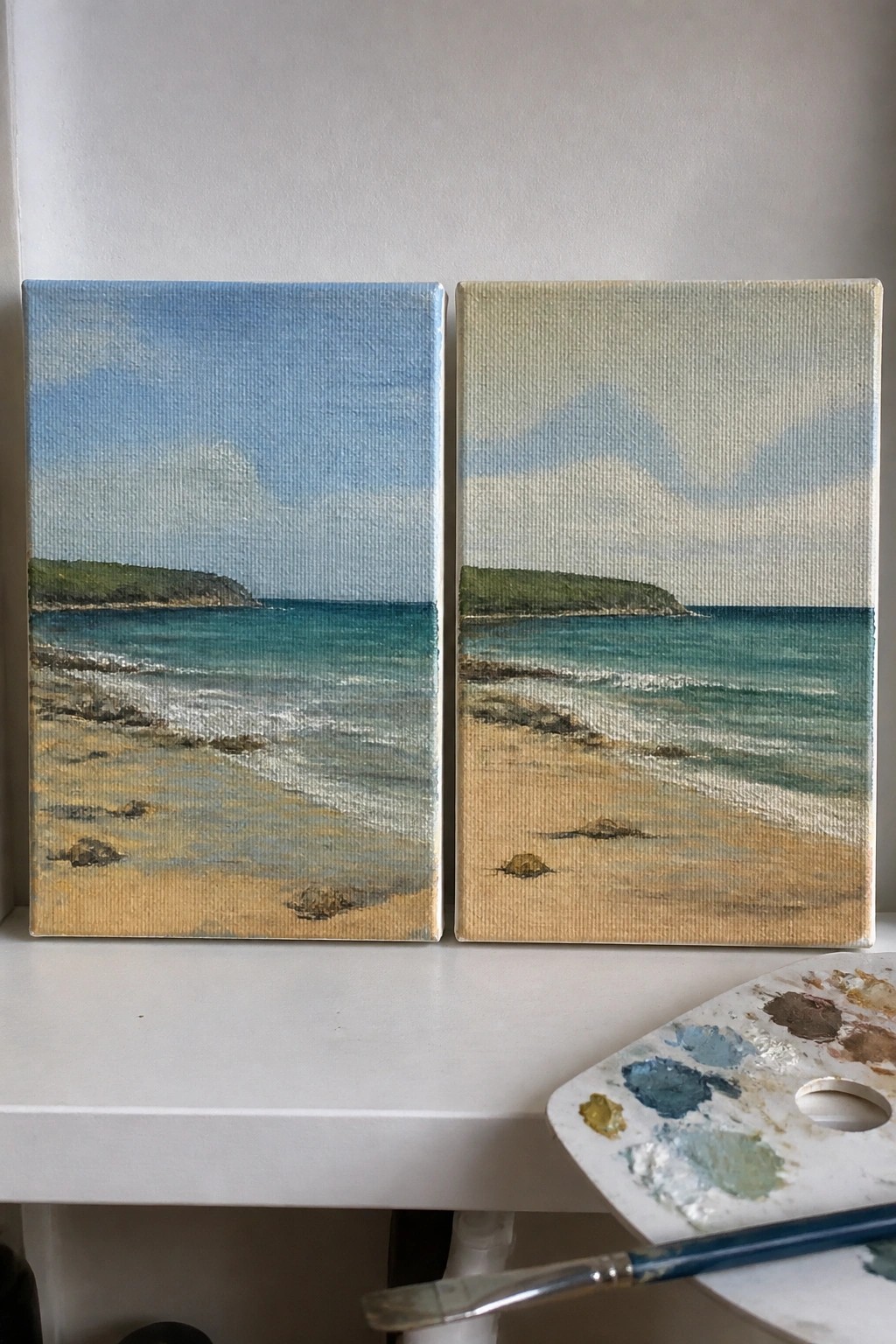 Two beach landscape paintings on canvas beside paint palette and brush