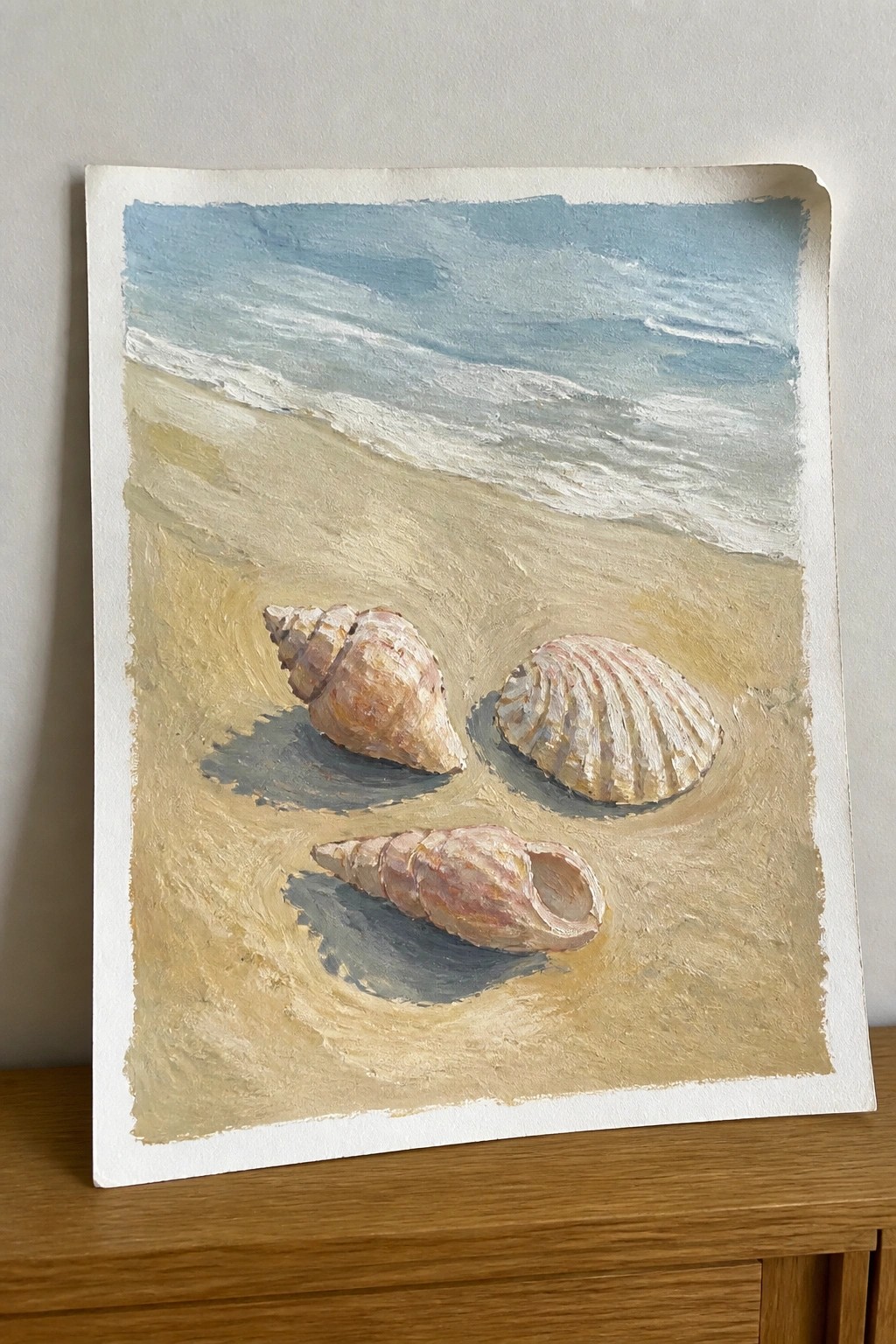 Three seashells on sandy beach with blue ocean waves in soft-toned painting.