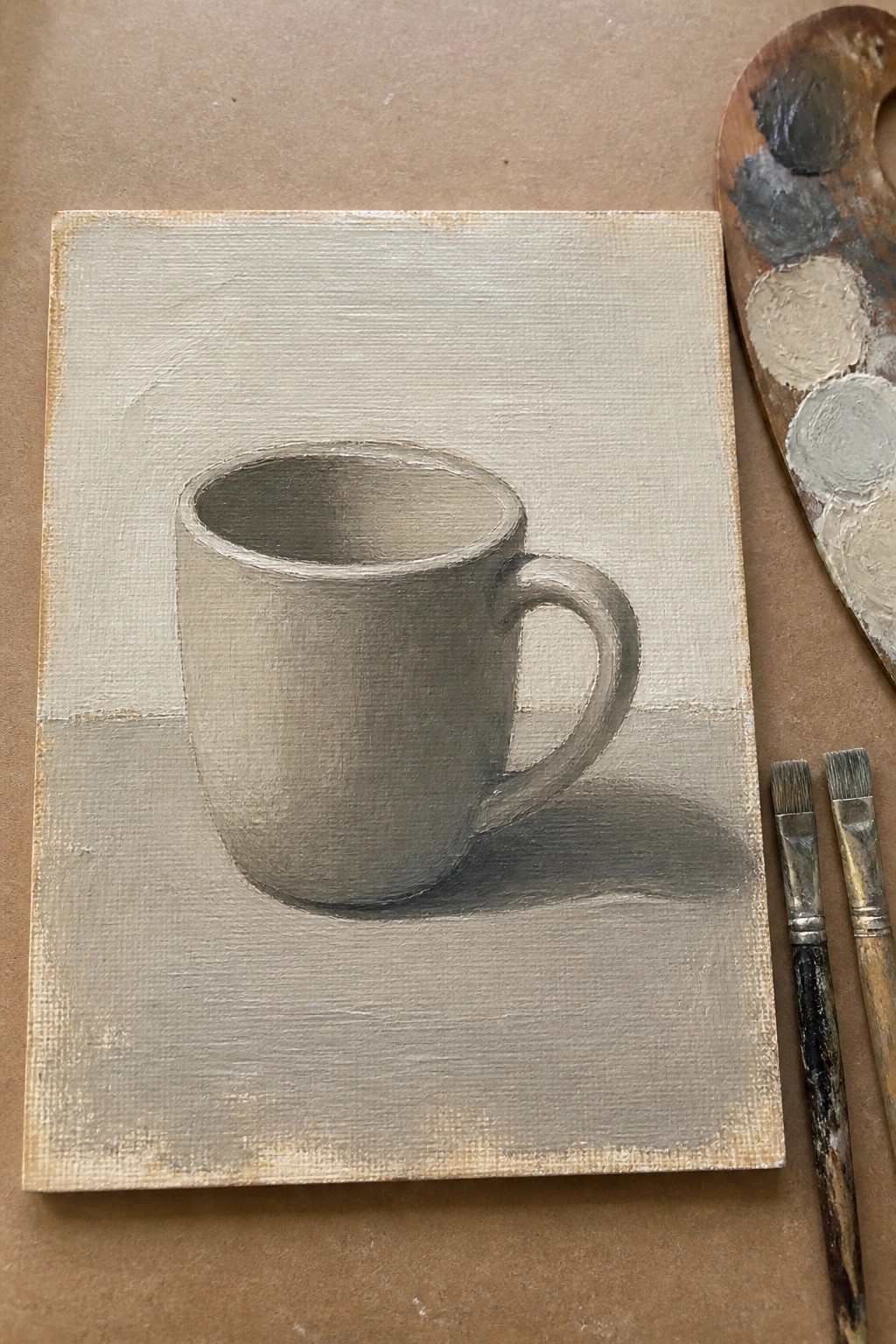 Monochrome painting of a gray mug with handle beside palette and brushes on canvas