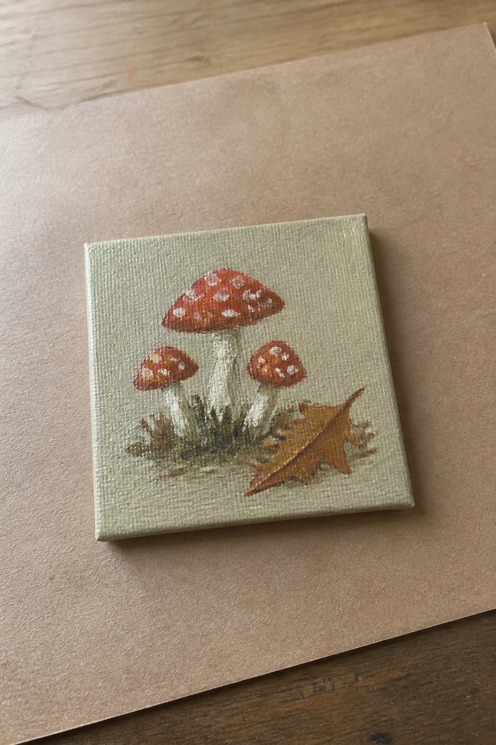 Small canvas painting of three red spotted mushrooms with a brown leaf on light green.