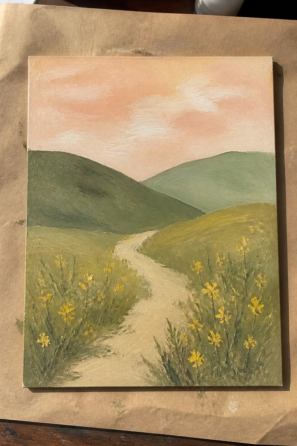 Winding path curves through green hills with yellow wildflowers under a peach sky.