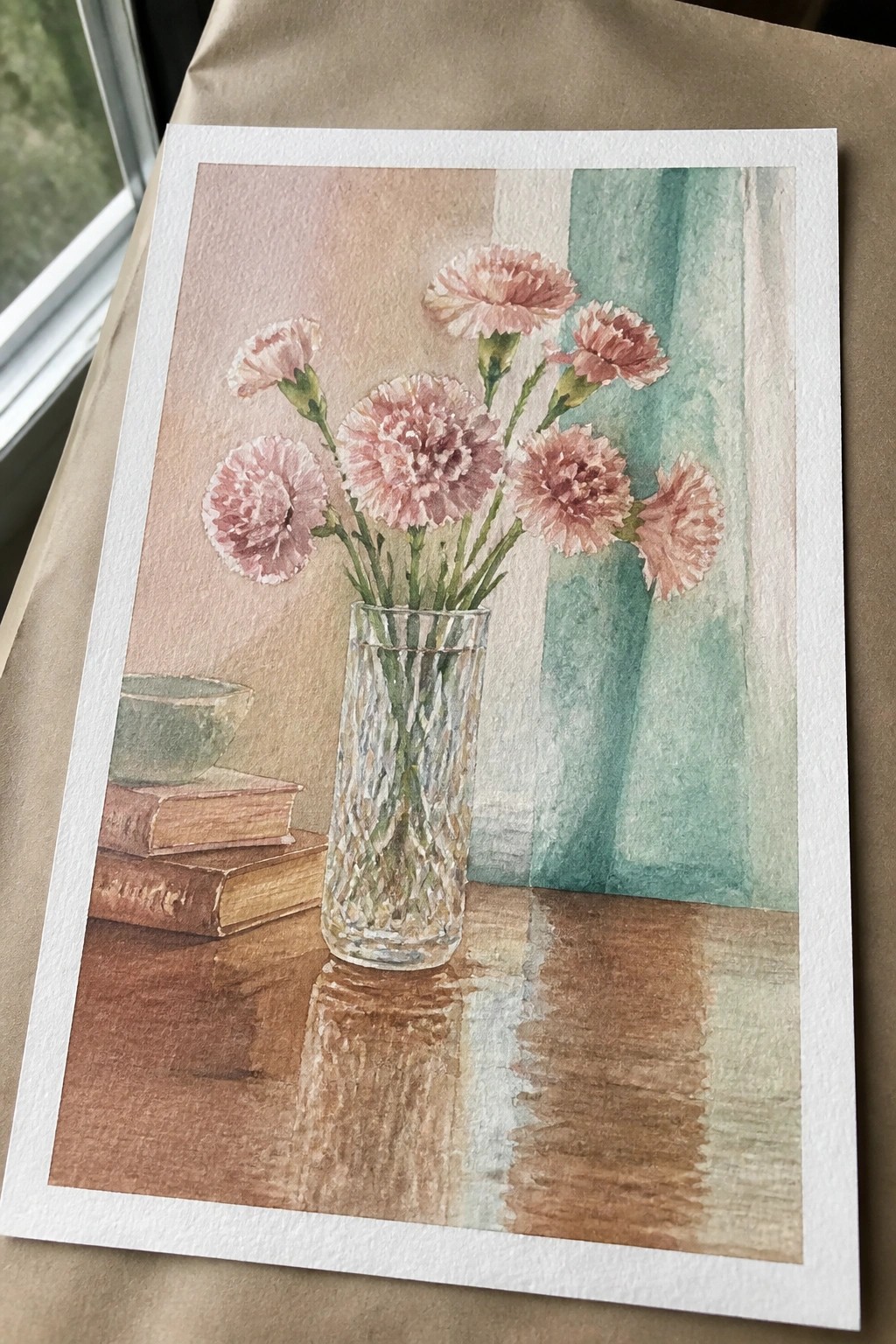 Watercolor of pink carnations in a glass vase on a wooden table with books.