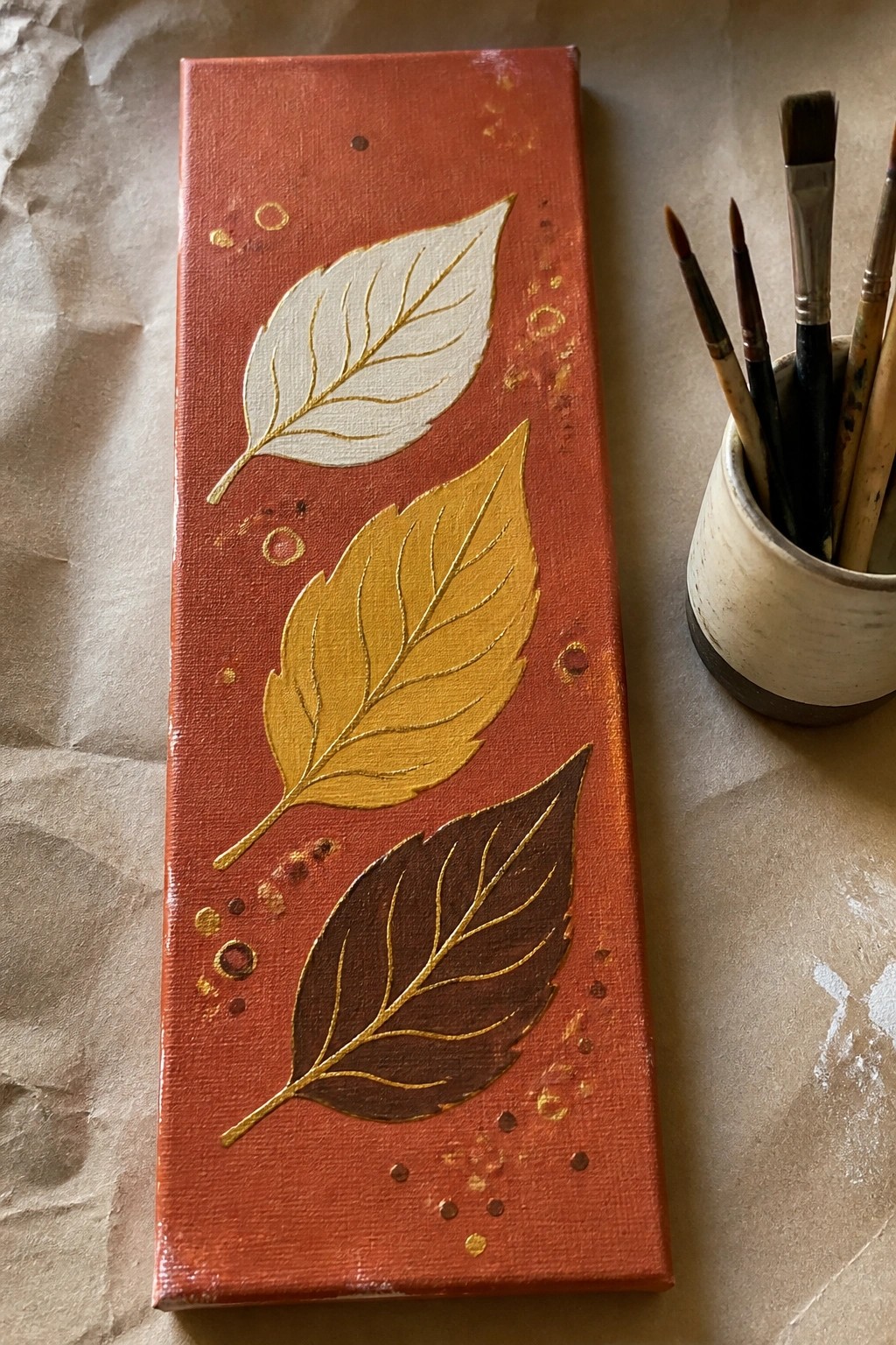 Red-orange canvas with three gold-veined leaves in white, yellow, and brown beside paintbrushes.