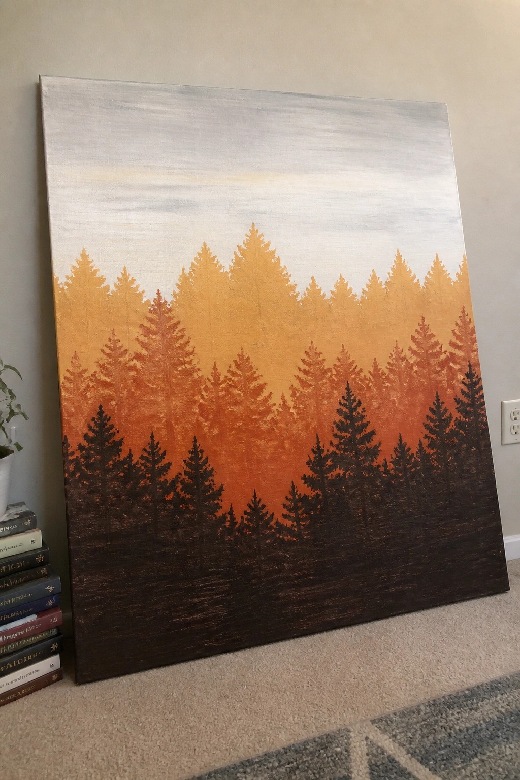 Large canvas painting of gradient orange pine forest leaning beside stack of books