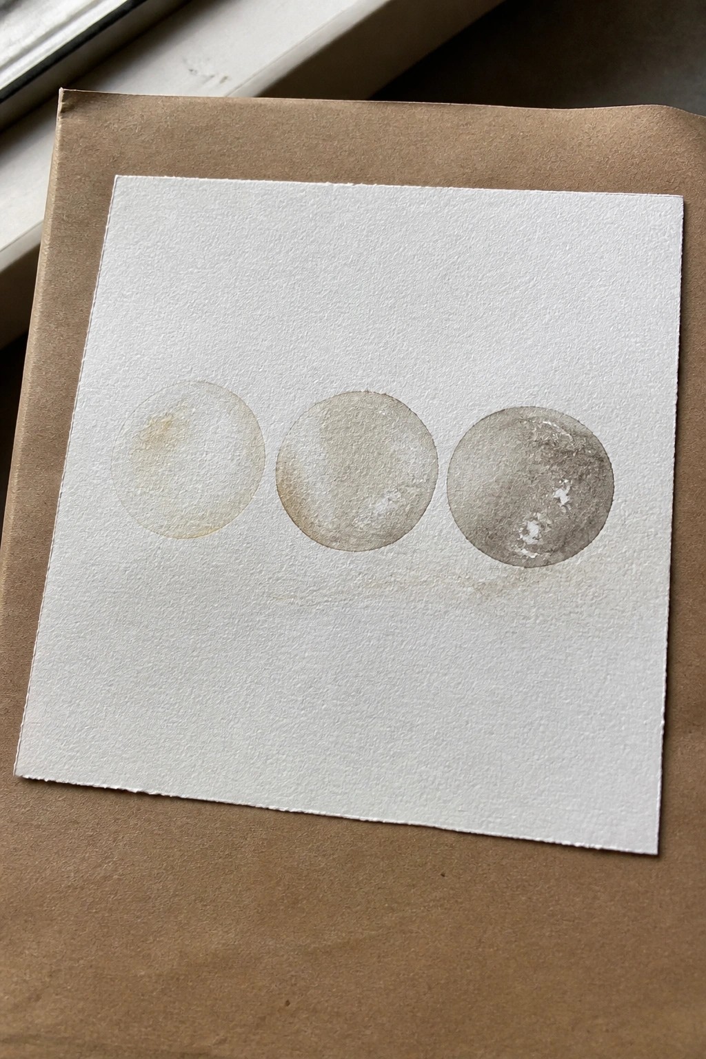 Three watercolor circles fading from light to dark on white paper over brown surface