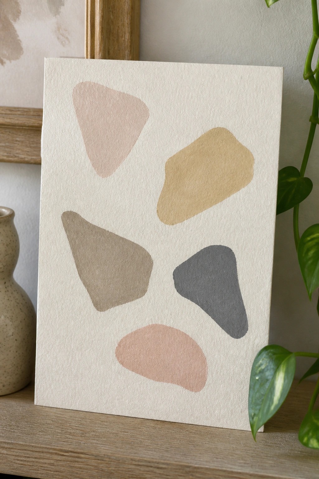 Abstract artwork with five irregular neutral shapes on textured white paper beside plant and vase.