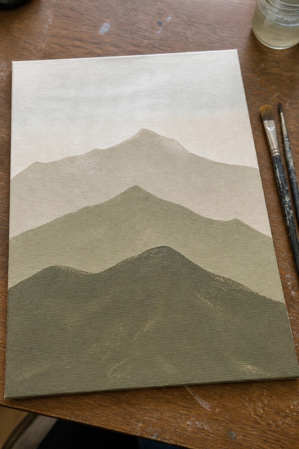Minimalist canvas painting of layered green mountains beside paintbrushes on wooden table.
