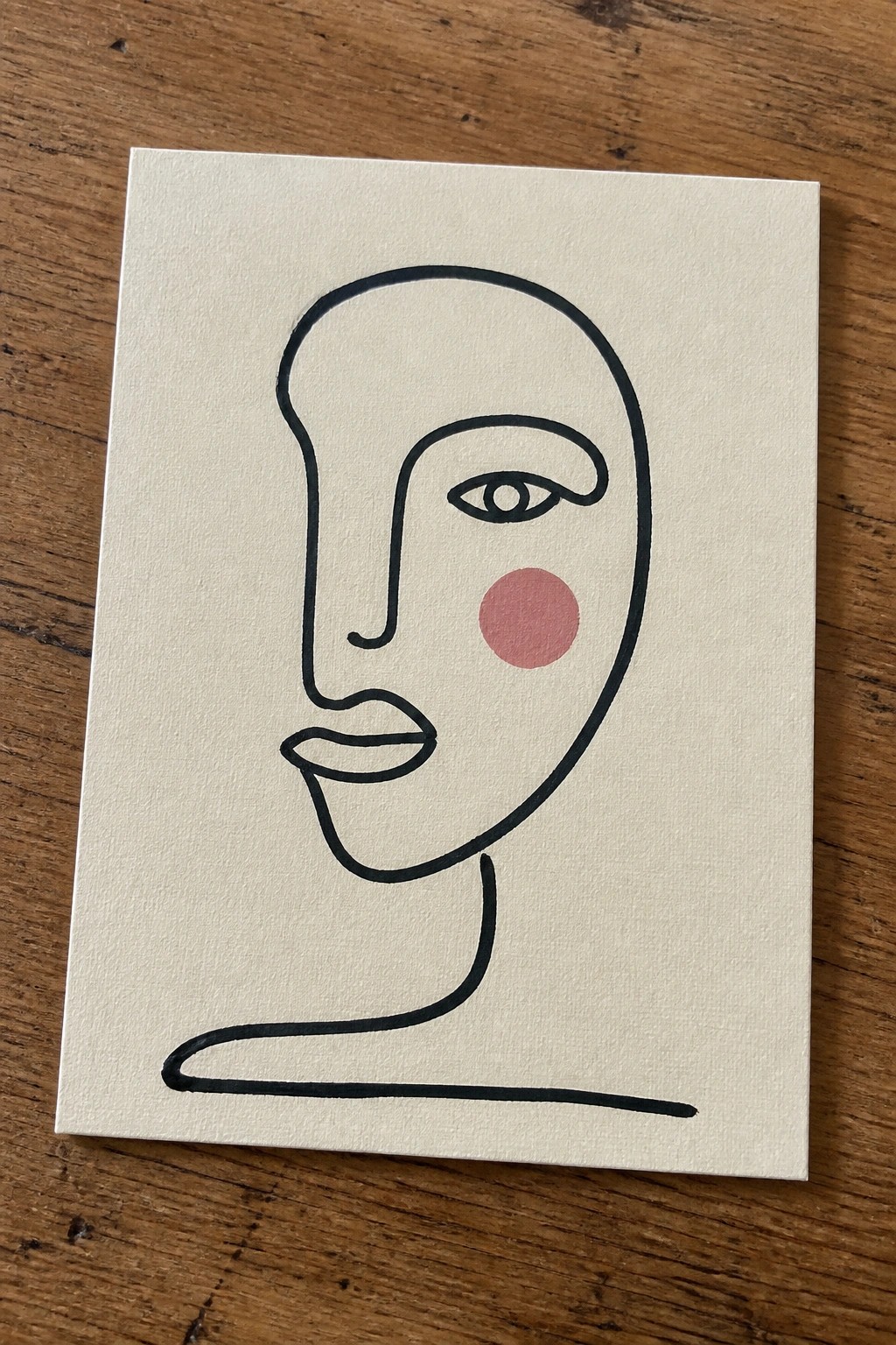 Minimalist line drawing of a face with pink cheek on white canvas over wood