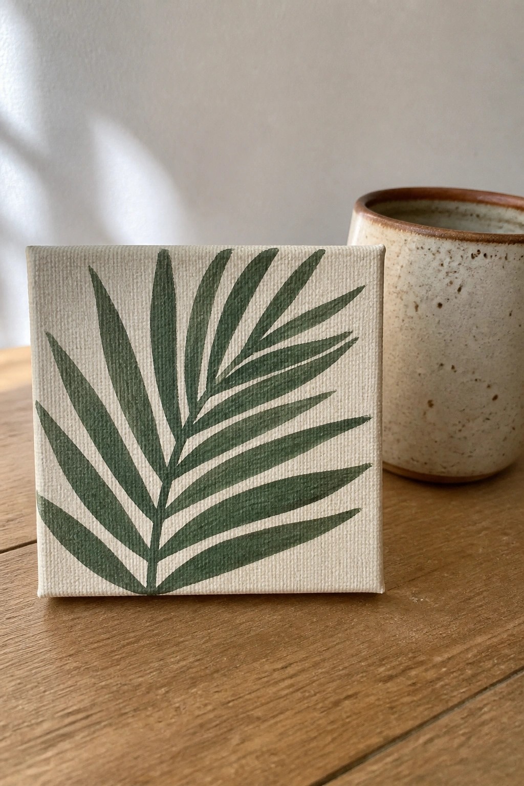Small canvas with green palm frond painting beside speckled ceramic cup on wood table
