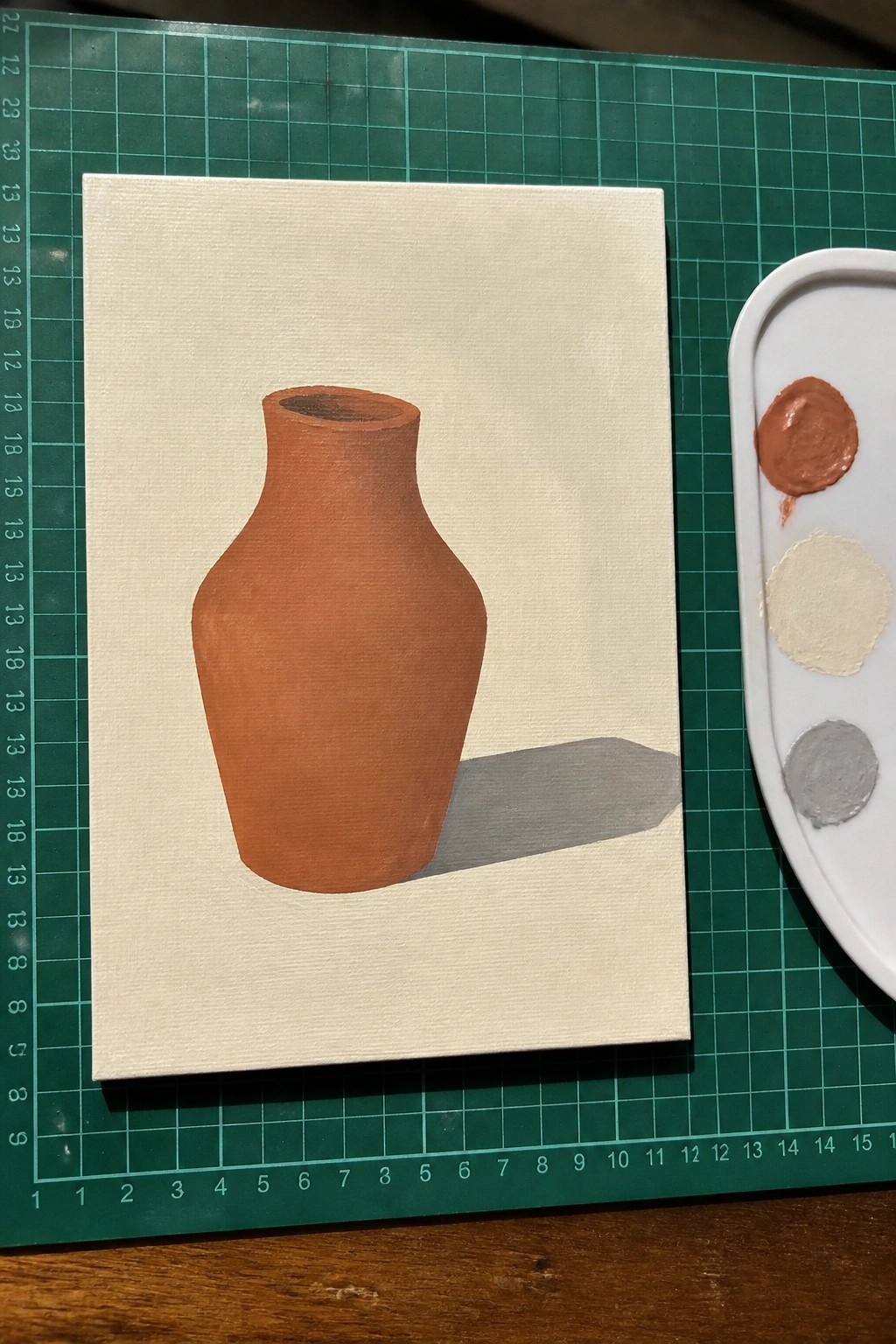 Acrylic painting of a terracotta vase with a gray shadow on a light canvas next to a paint palette on a green mat.