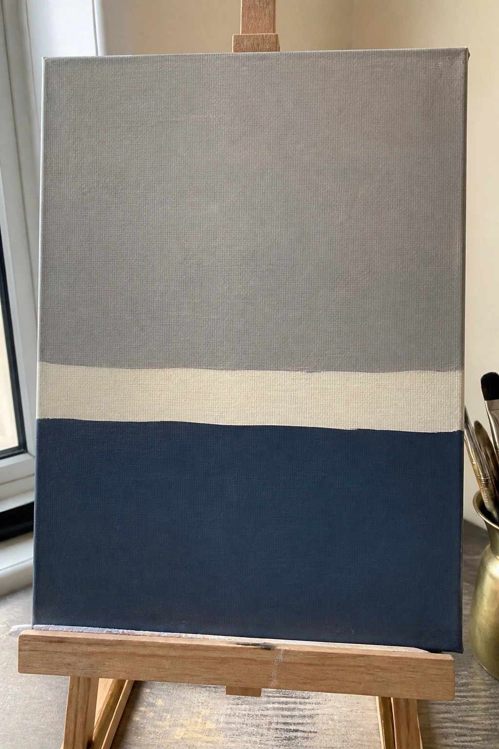 Canvas on easel with horizontal gray, white, and dark blue painted stripes, brushes nearby