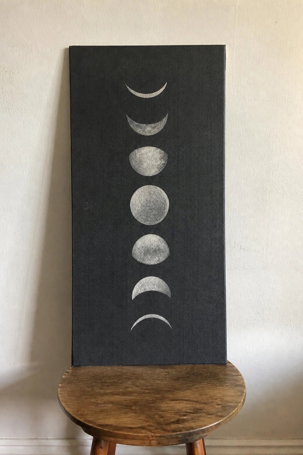 Black canvas with white moon phases on round wooden stool against wall