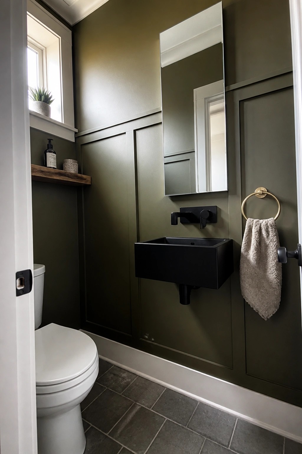 A small bathroom with deep green paneled walls, a black floating sink, and brass accents.