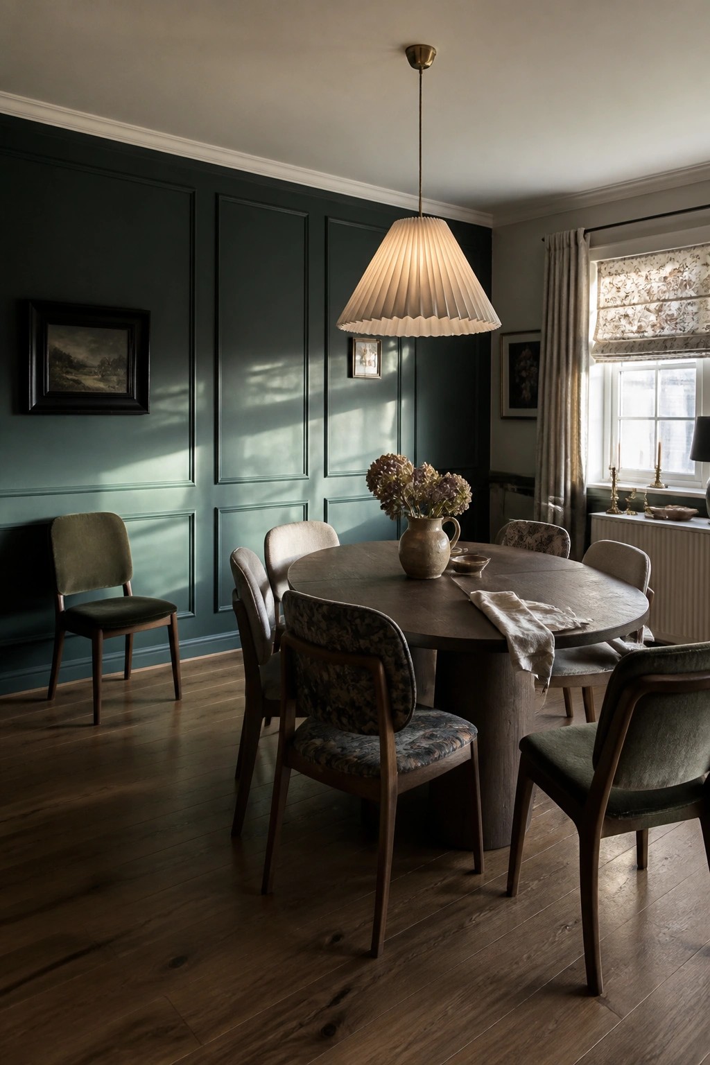 Round wooden dining table with mixed chairs beneath pleated pendant light in dark green room.
