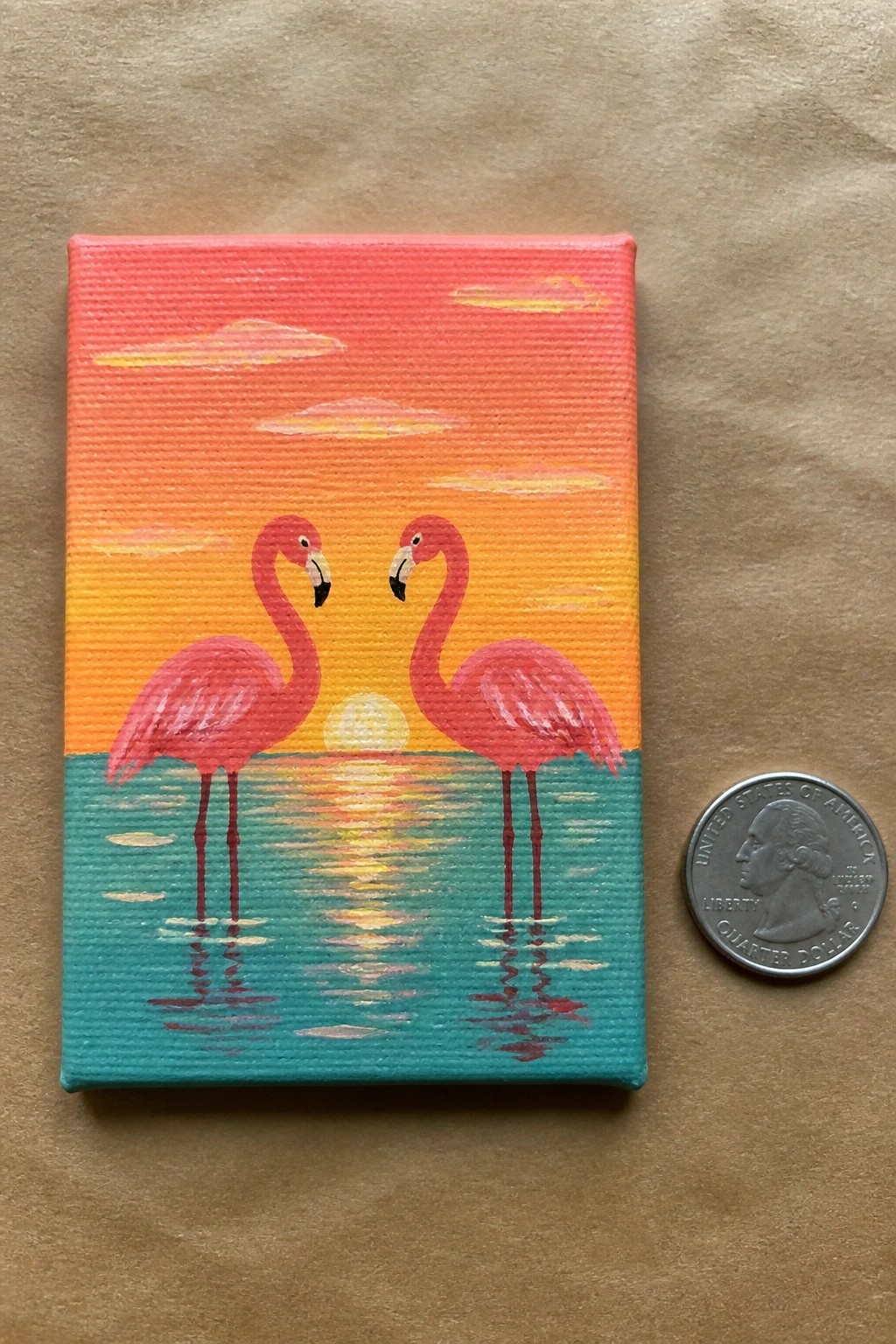 A small acrylic painting of two pink flamingos standing in turquoise water under an orange and yellow sunset sky, with a quarter coin next to it for scale.