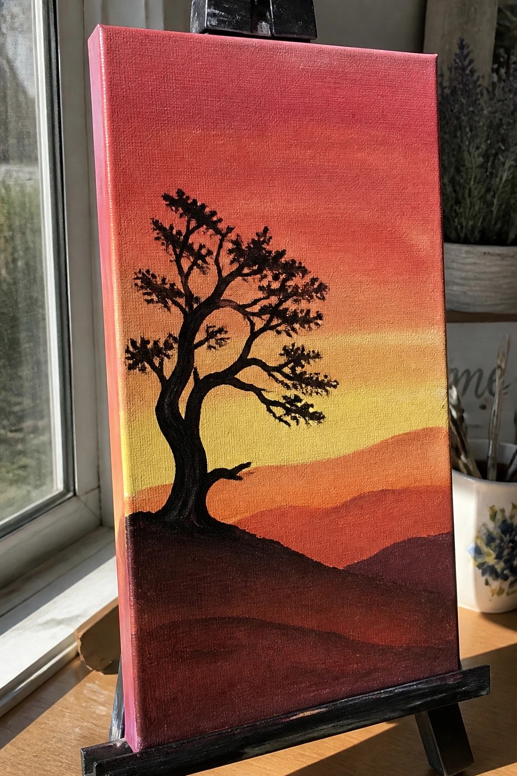 Silhouetted tree against orange-red-yellow gradient sunset sky with layered hills on easel