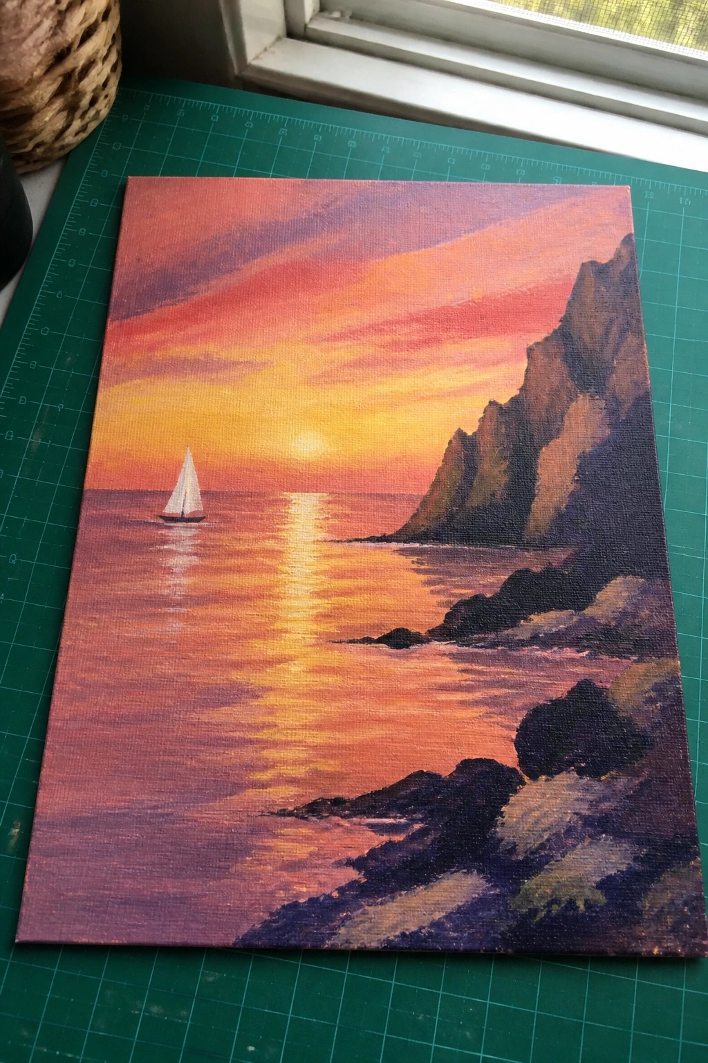 Painting of sailboat on orange sunset ocean beside steep rocky cliffs on green mat.