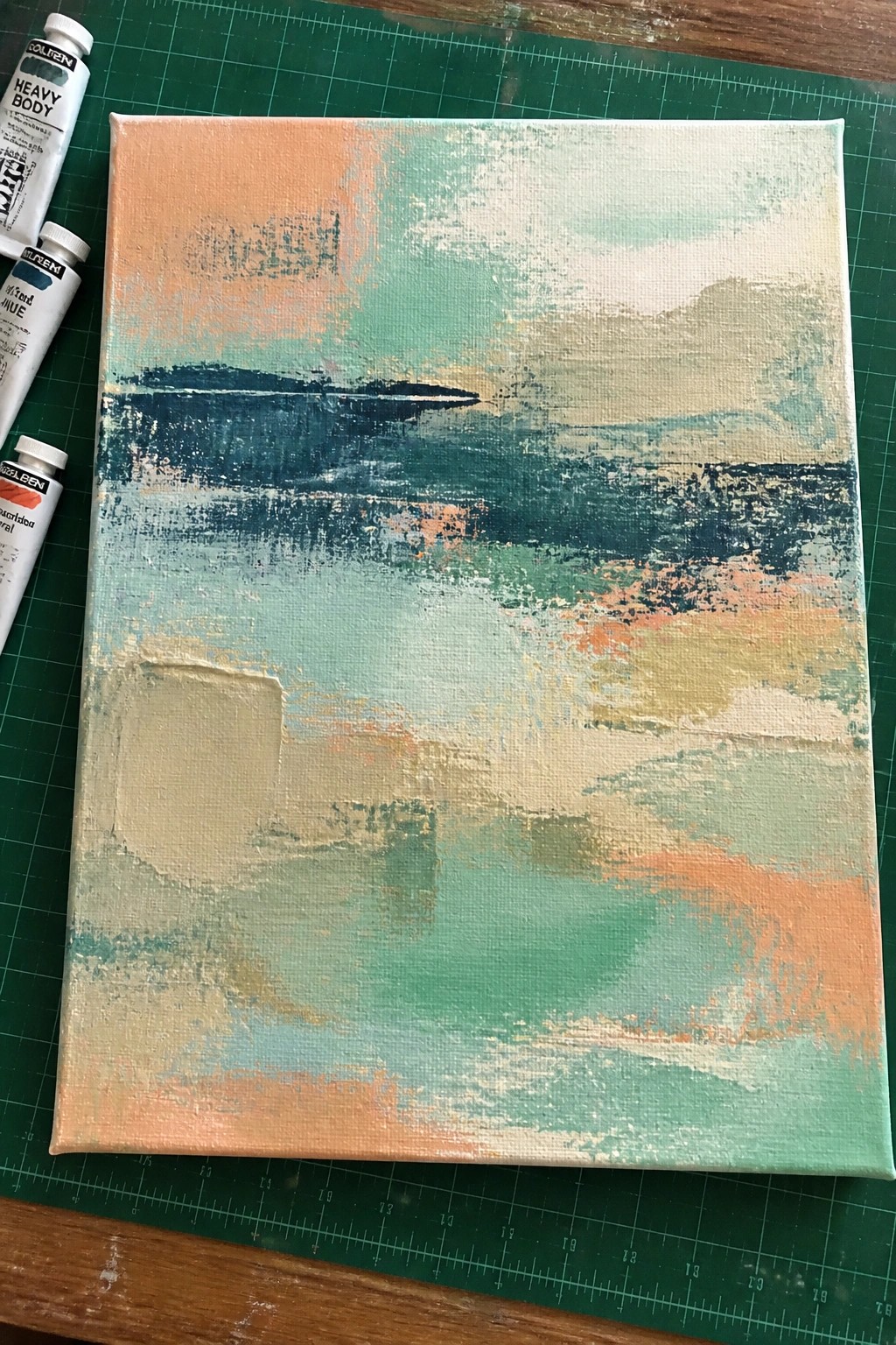 Abstract painting with teal, orange, blue, and green brushstrokes on canvas beside paint tubes.