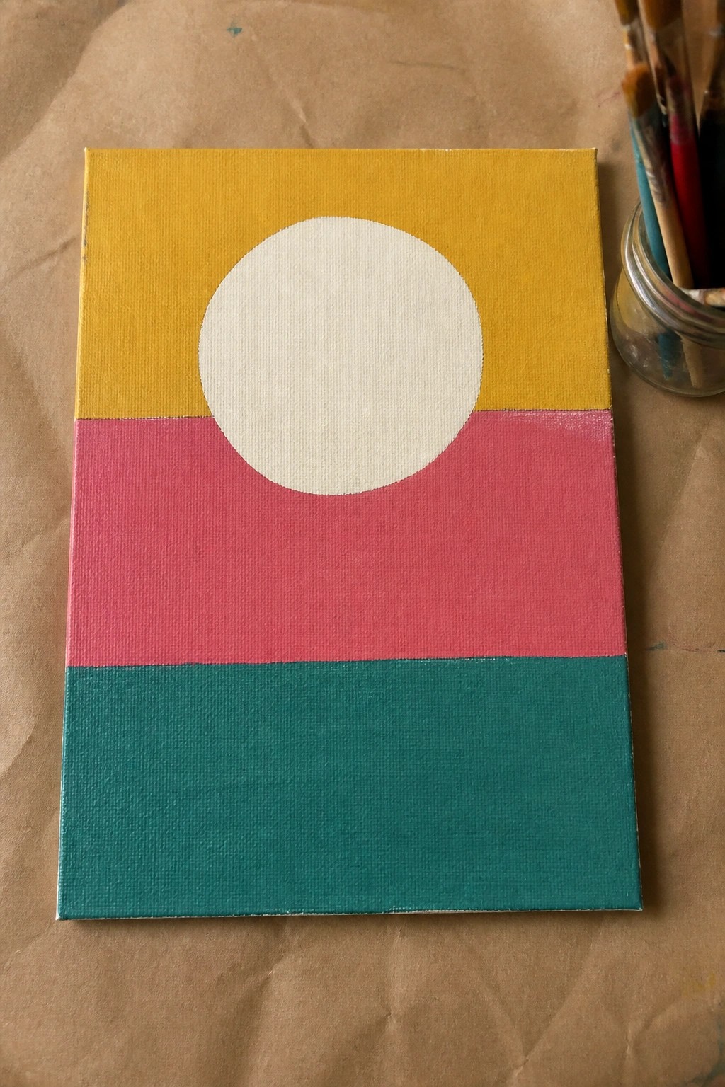 Abstract painting with yellow top, pink middle, teal bottom, white circle beside paintbrushes.
