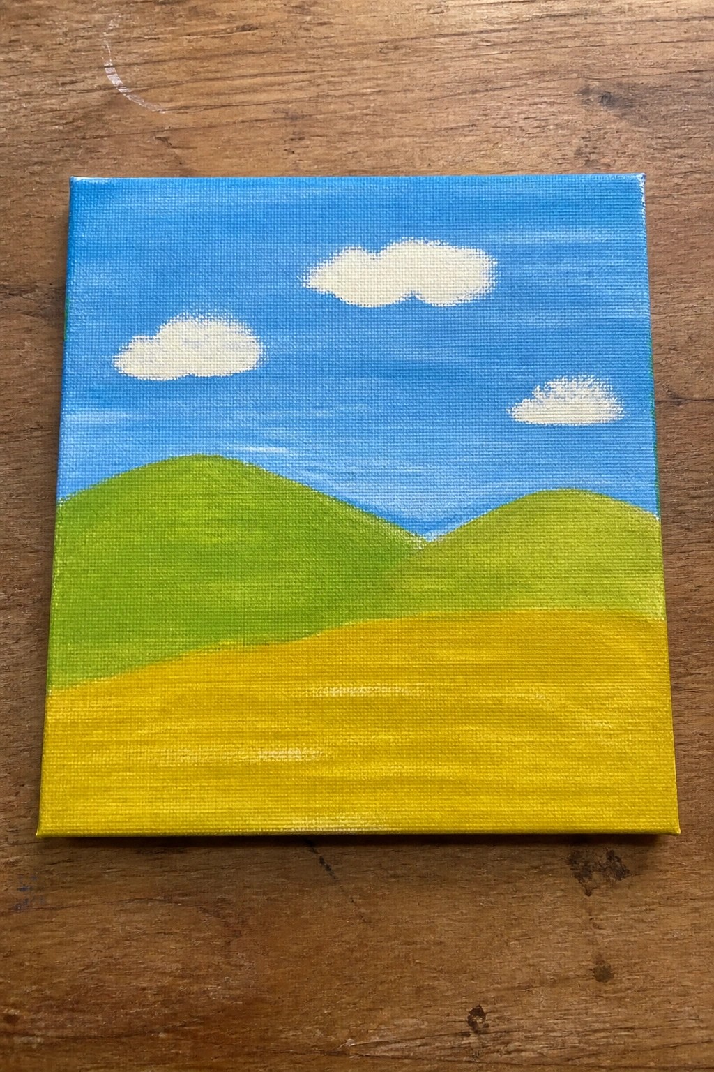Small canvas with blue sky, white clouds, green hills, and yellow field on wood.