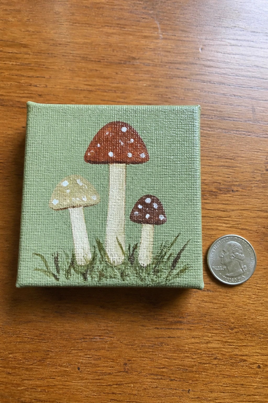Tiny painting of three spotted mushrooms on green canvas beside a quarter.