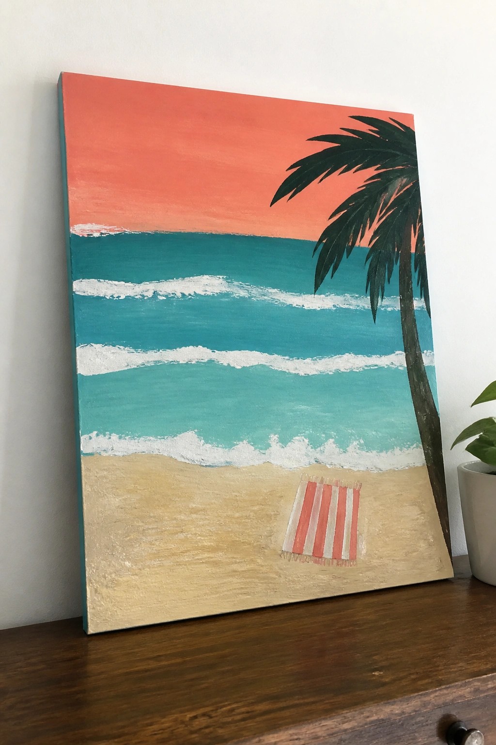 A rectangular acrylic painting of a beach at sunset featuring an orange sky, teal ocean with white waves, sandy shore, a dark green palm tree, and a red striped towel on the sand.
