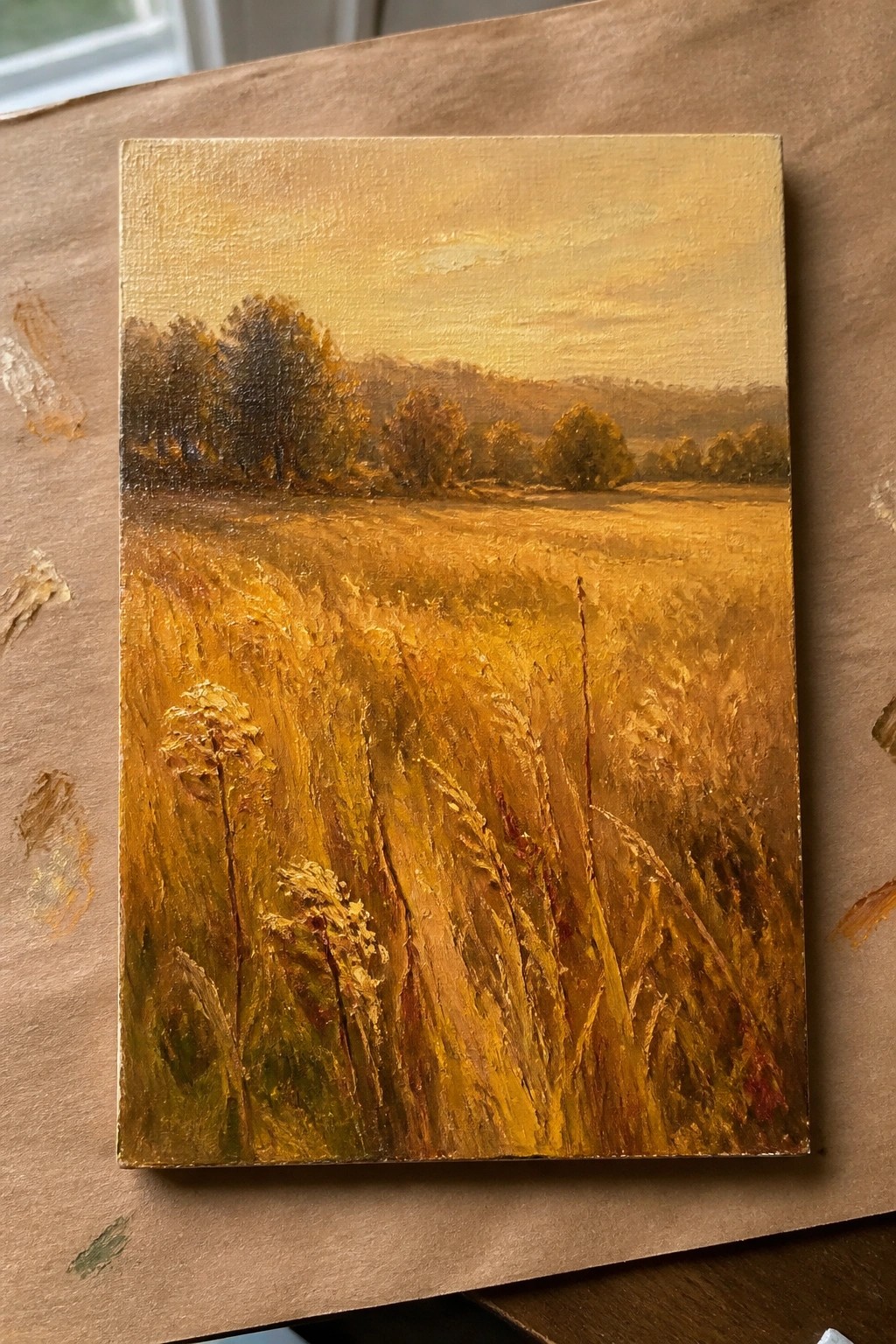 Oil painting of vast golden wheat field with tall grasses, distant trees, and hills at sunset.