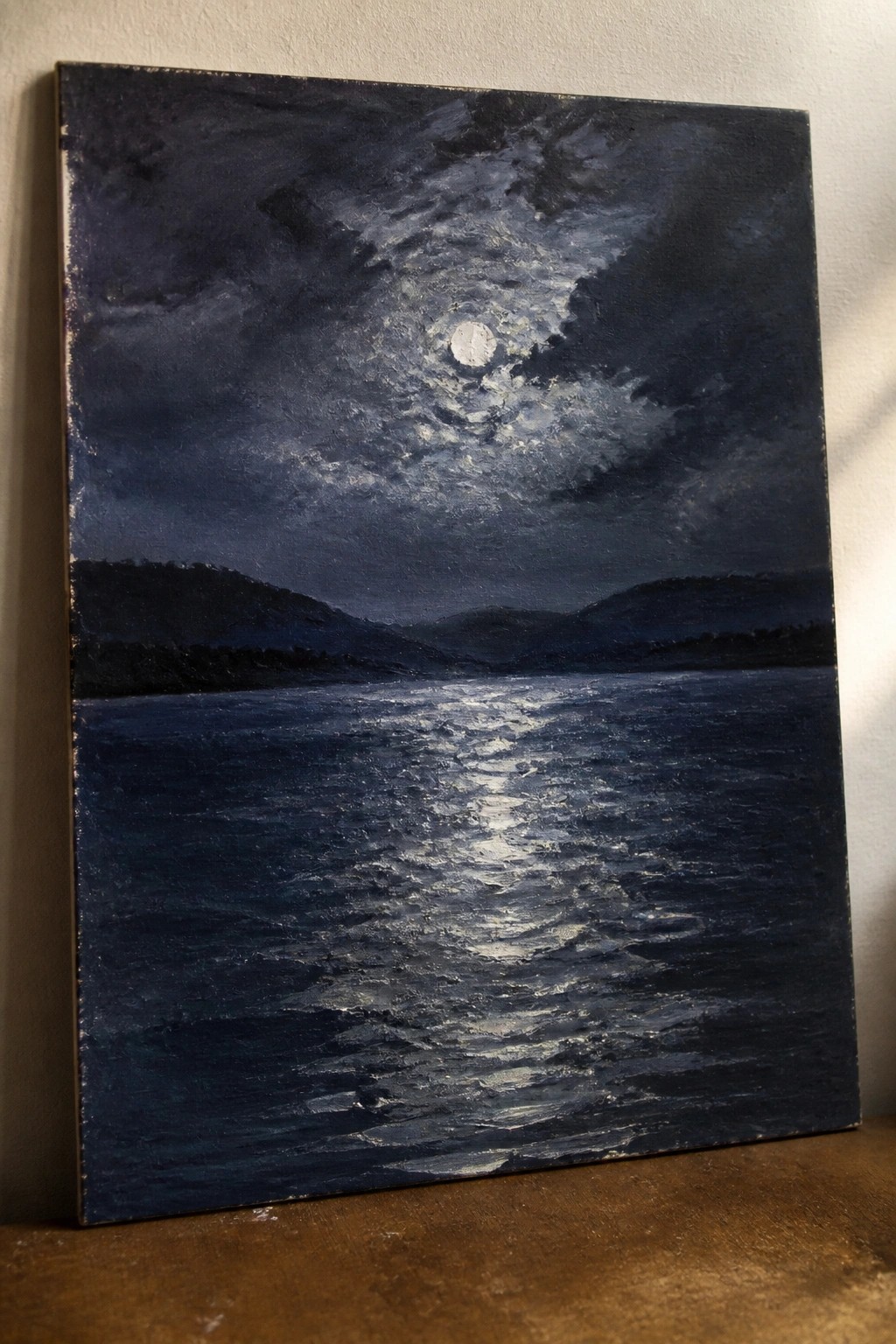 Oil painting of full moon reflecting on rippled dark water under cloudy night sky with hills.