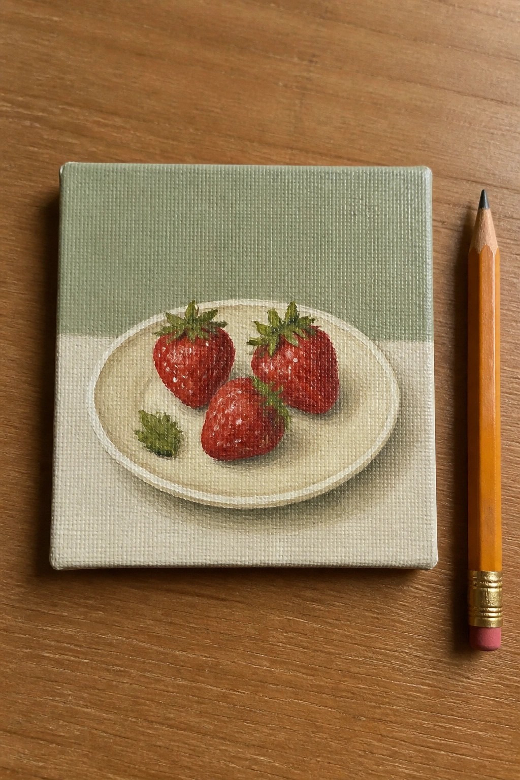 Small canvas painting of three strawberries on plate beside yellow pencil on wood