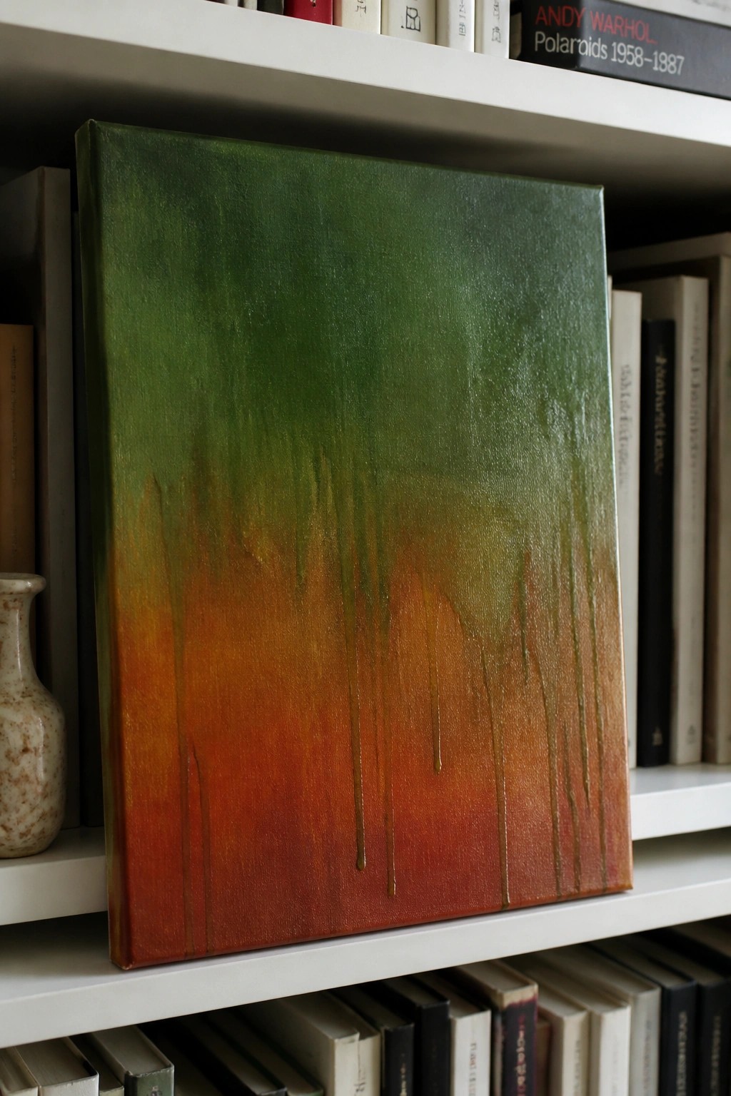 Abstract painting with green-to-orange gradient and vertical drips on bookshelf among books