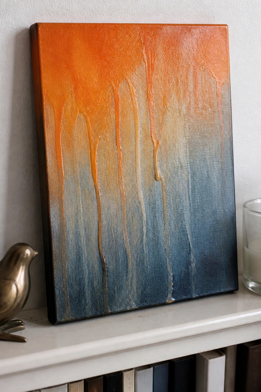 Abstract painting with orange-to-blue drips on white shelf beside bronze bird figurine.