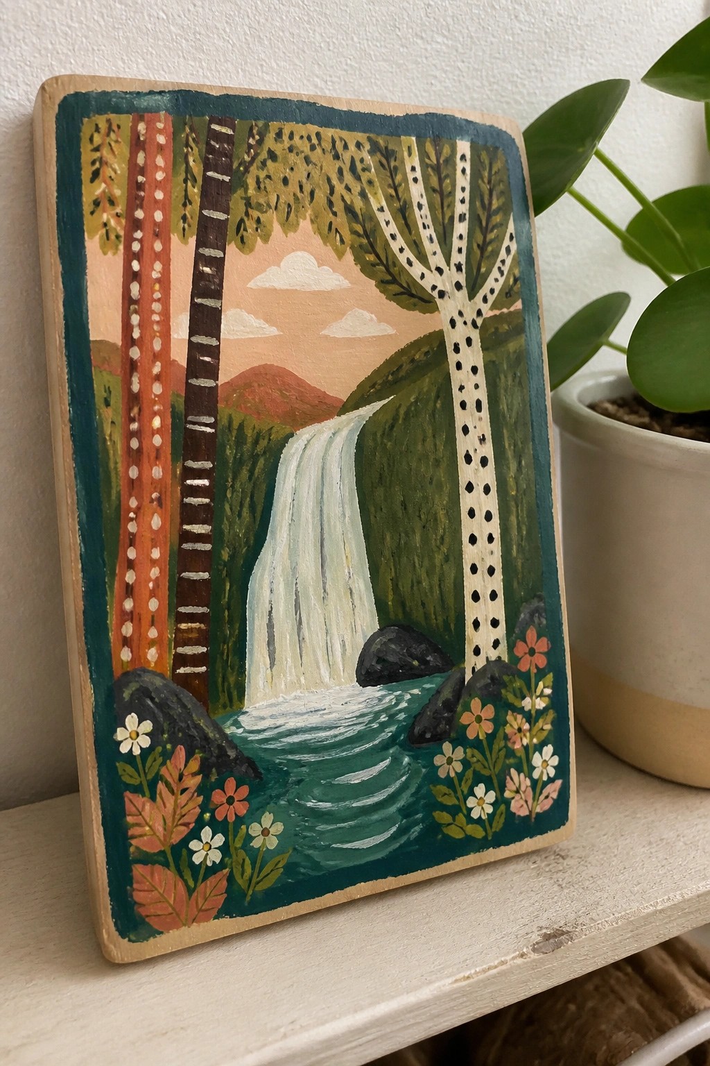 A painted wooden plaque showing a waterfall between trees with flowers in the foreground and a potted plant nearby.